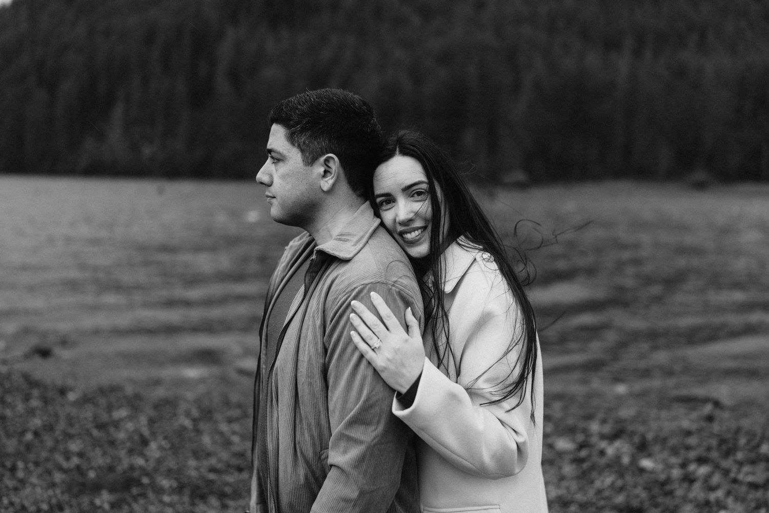 A happy couple embraces while standing near a lake with mountains in the background.