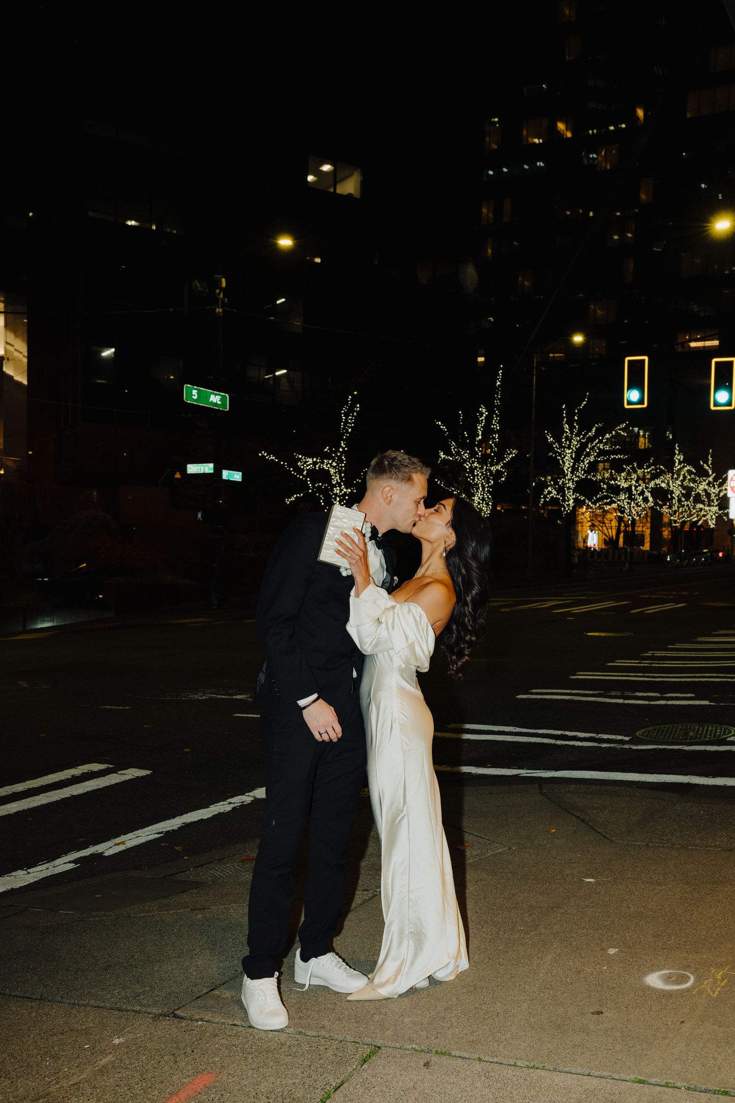 A couple shares a romantic nighttime kiss on a city street decorated with illuminated trees and holiday lights.