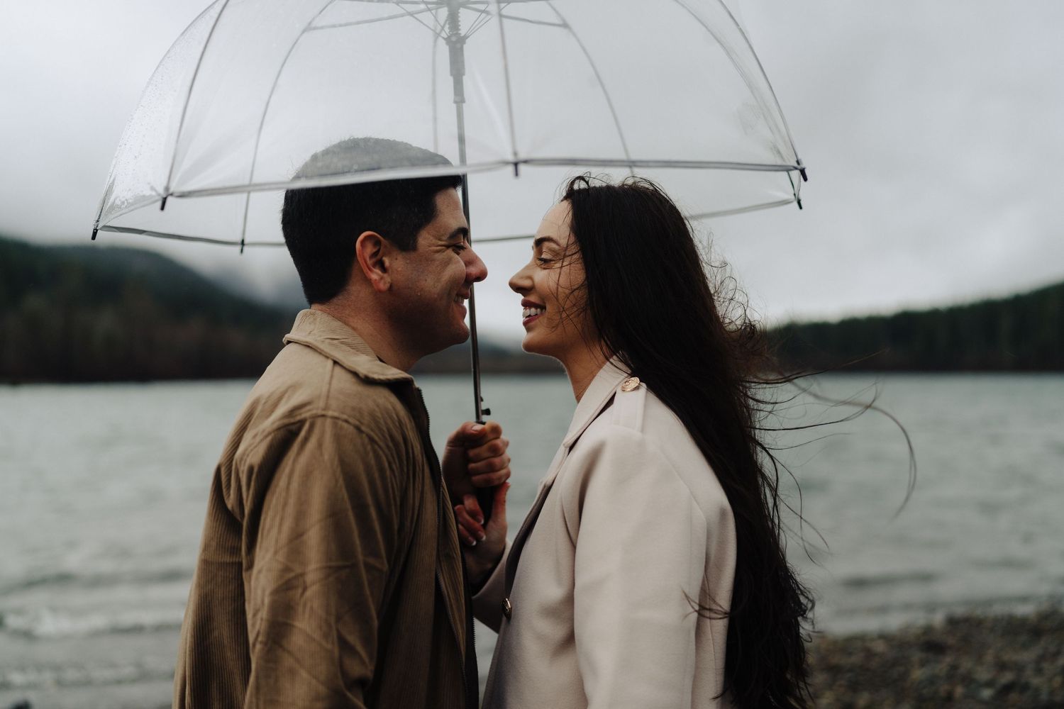A couple shares an intimate moment under a clear umbrella beside a lake on an overcast day.