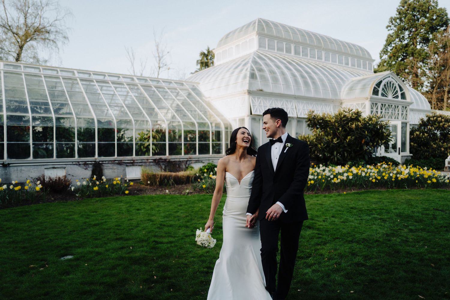A wedding couple posing outside a grand glass conservatory with beautiful landscaping.