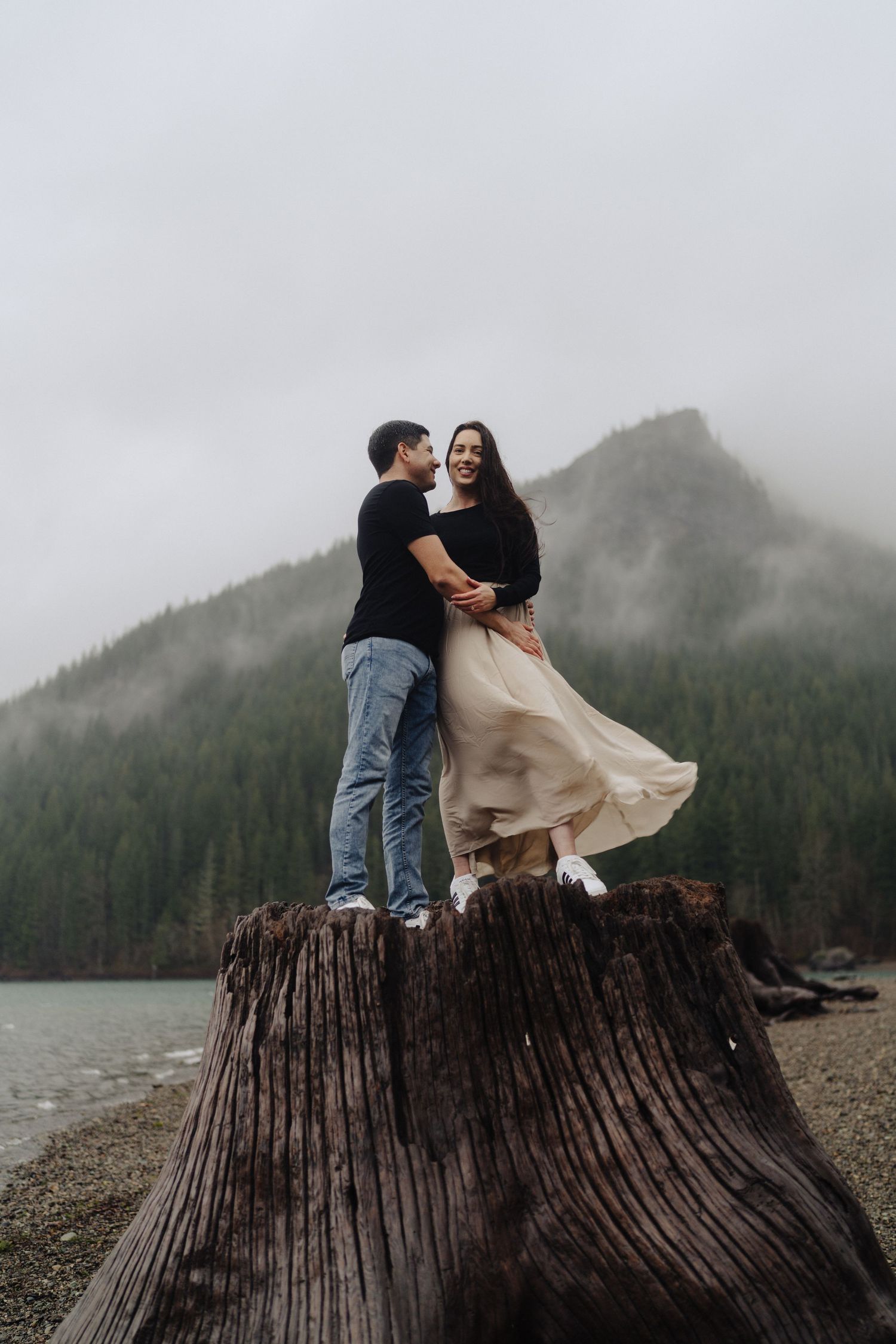 Romantic couple stands on large tree stump by mountain lake with dress flowing in the wind.