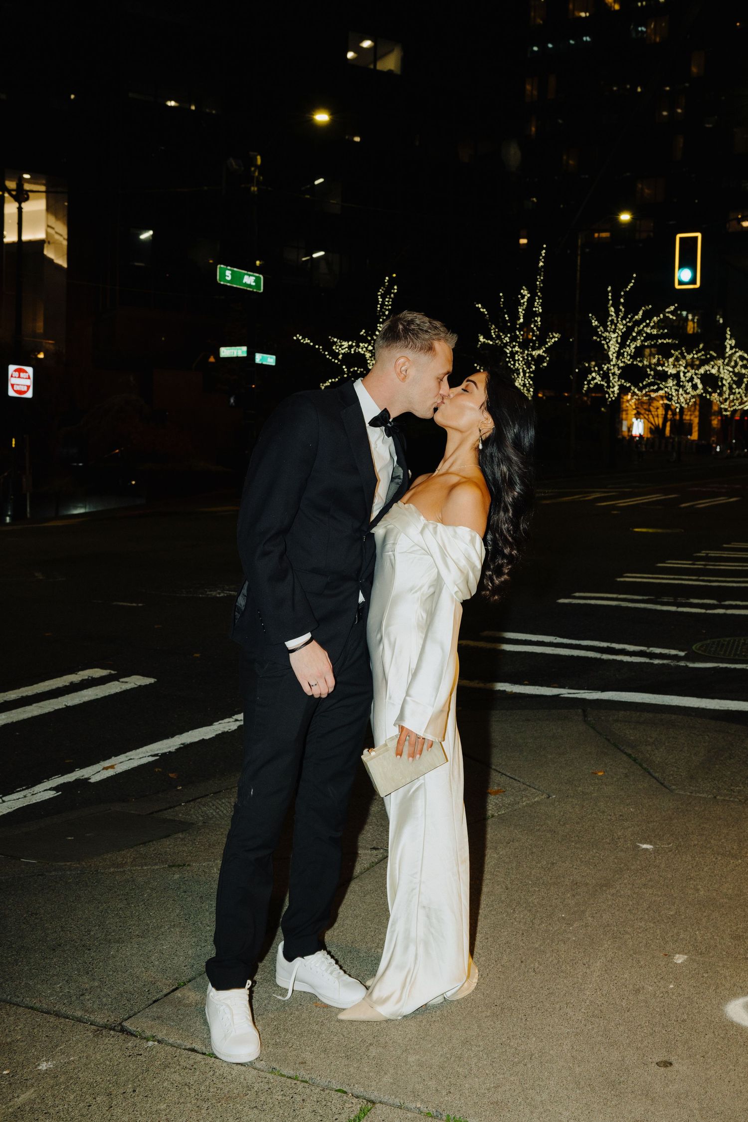 Romantic nighttime street scene of newlyweds sharing a kiss under city lights.