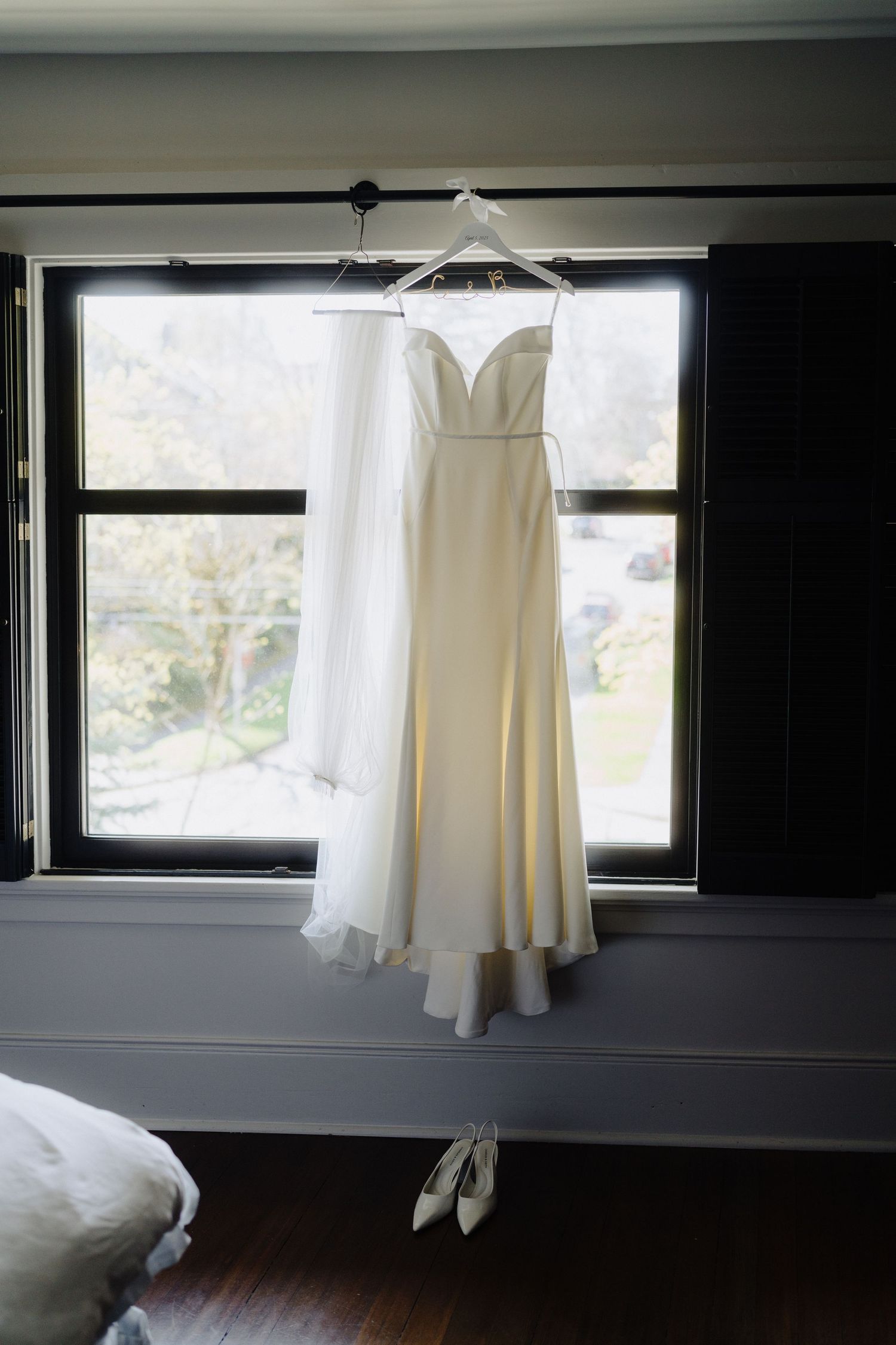Elegant white wedding dress with sweetheart neckline hanging in window with ballet shoes below.