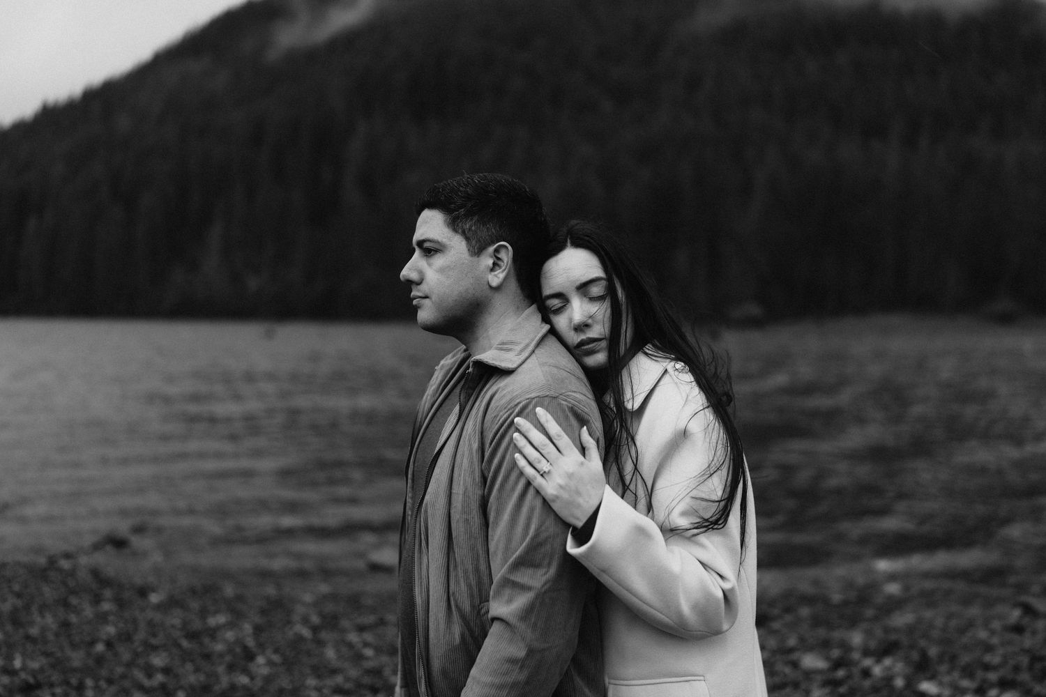 A couple stands together by a lake with mountains in background captured in dramatic black and white photography.