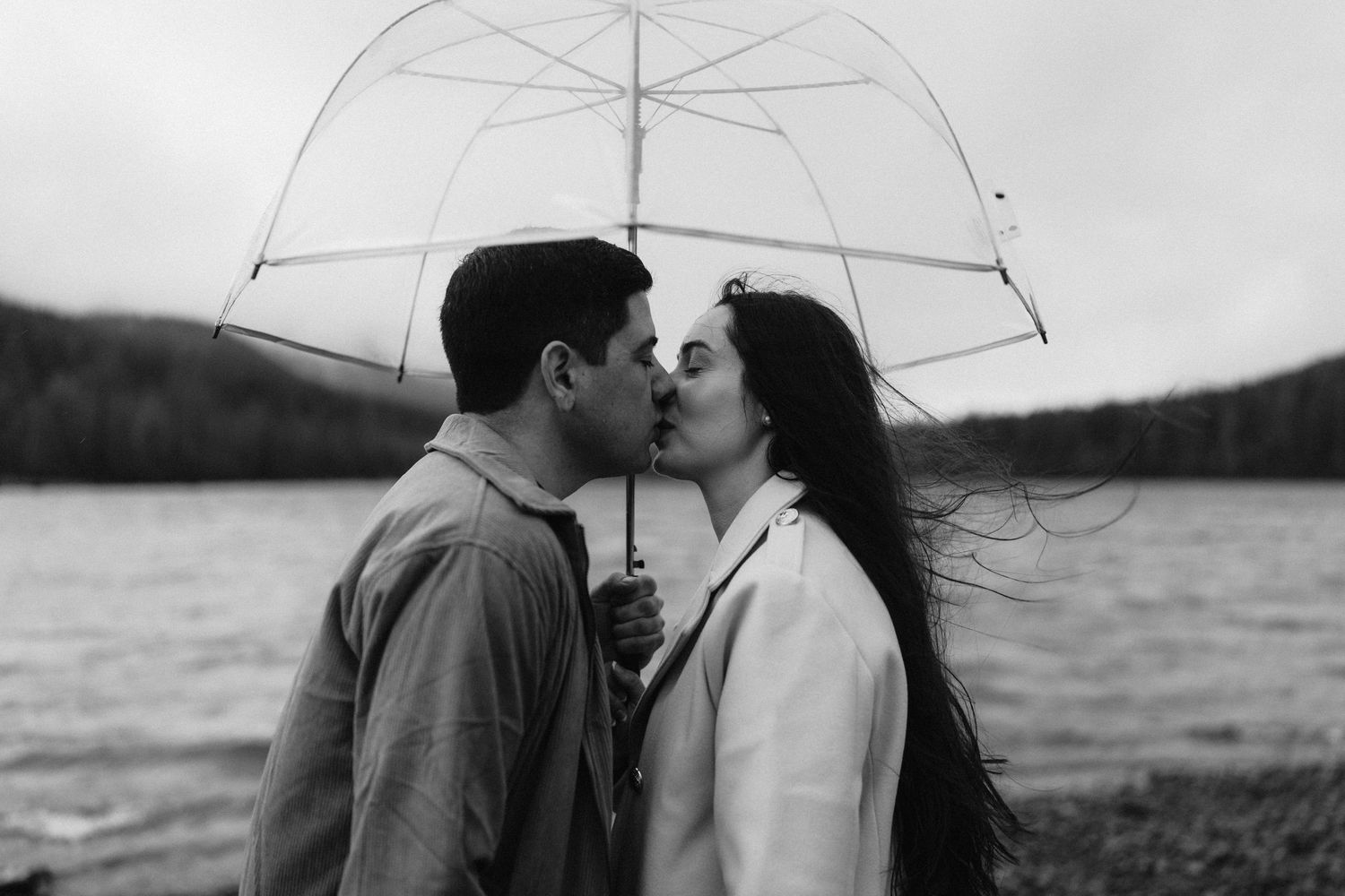 A romantic couple shares a kiss under a clear umbrella by a lake on an overcast day.