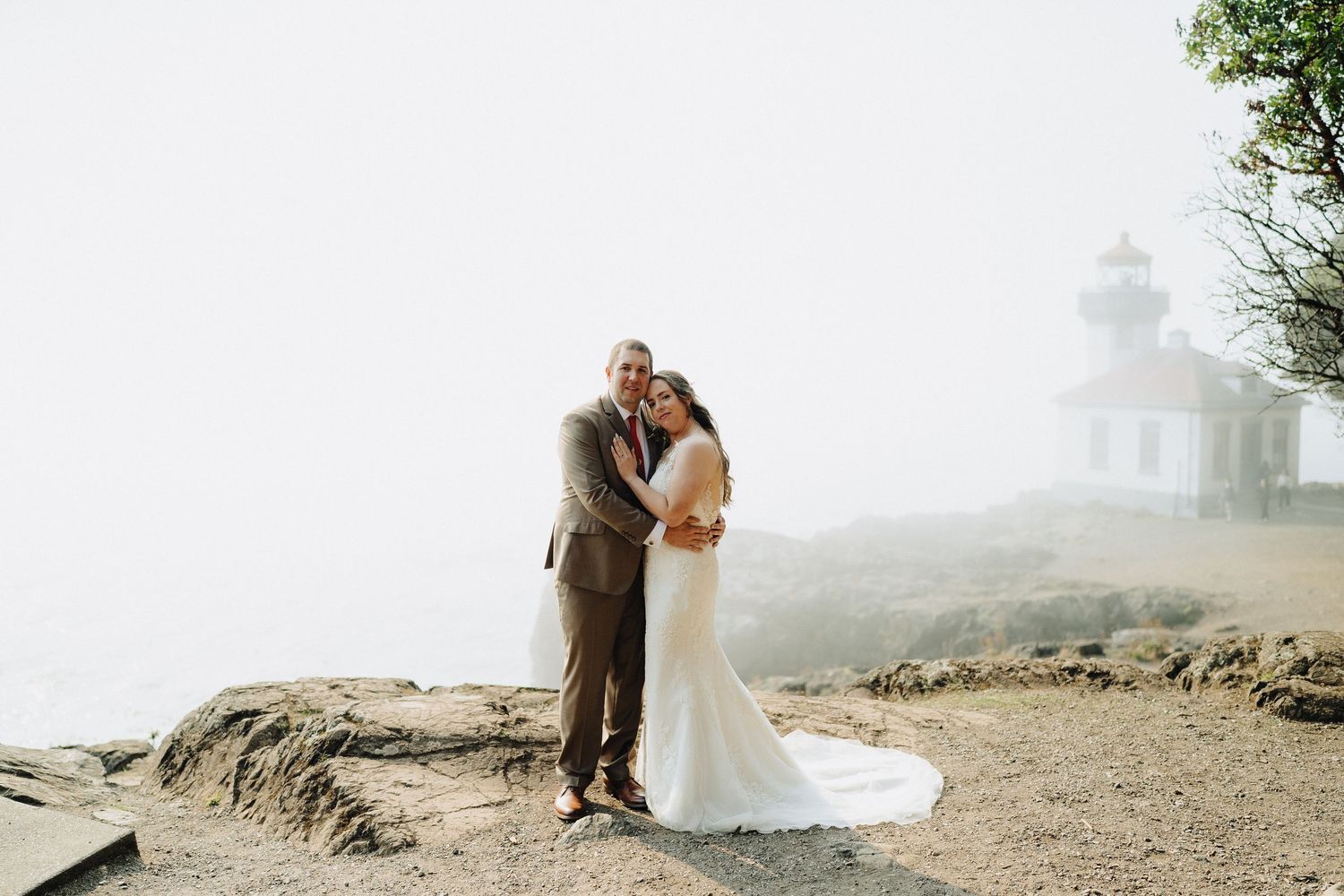 A couple embraces on a misty cliff overlooking the ocean with a lighthouse silhouette in the background.