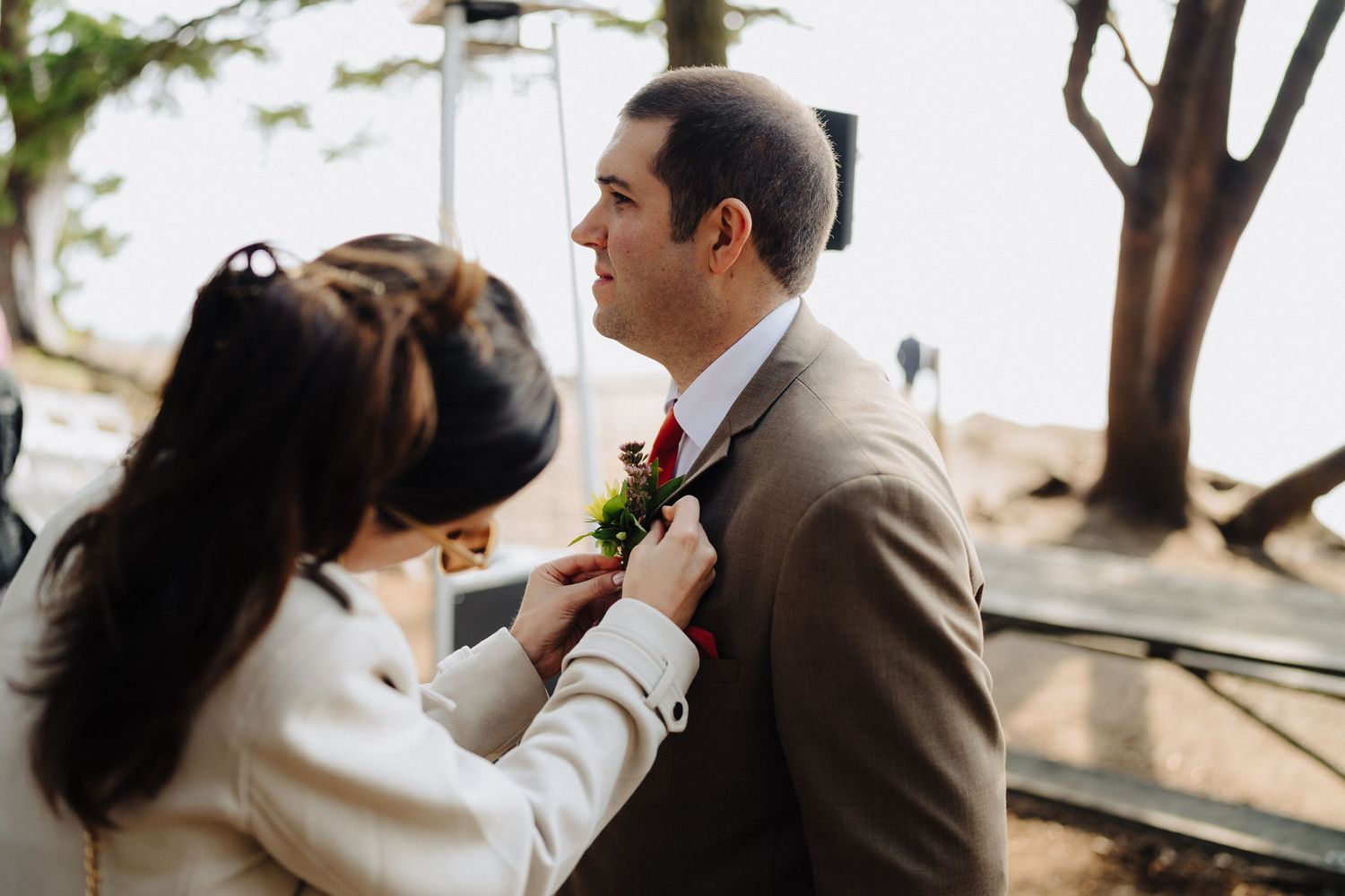 A couple shares a tender moment during a wedding ceremony as a red boutonniere is pinned to a tan suit jacket.