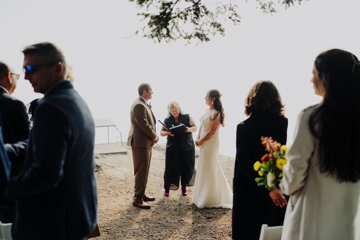 An intimate coastal wedding ceremony takes place on a cliff edge overlooking the ocean on a misty day.