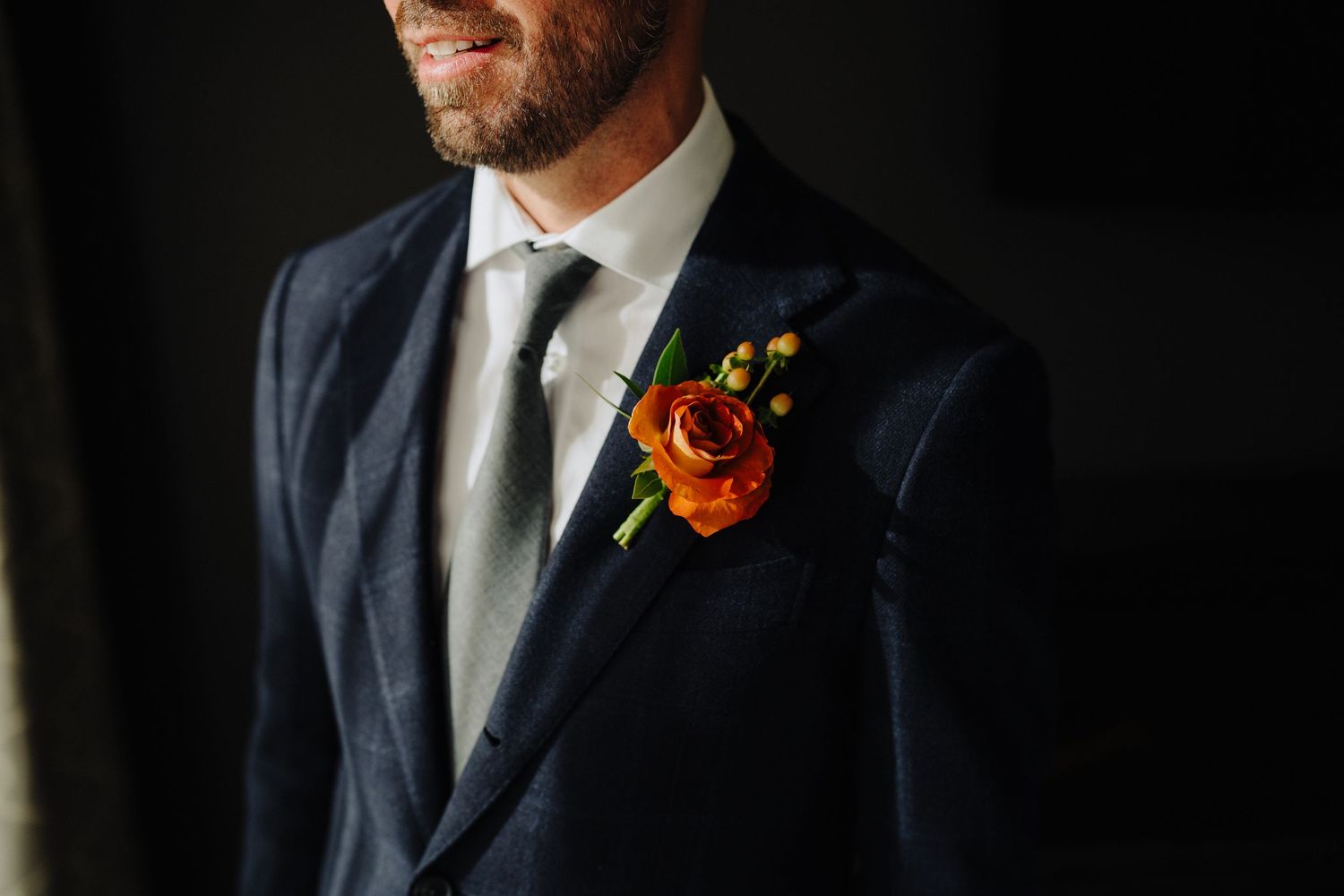 Close up of wedding boutonniere with orange rose on navy suit lapel.