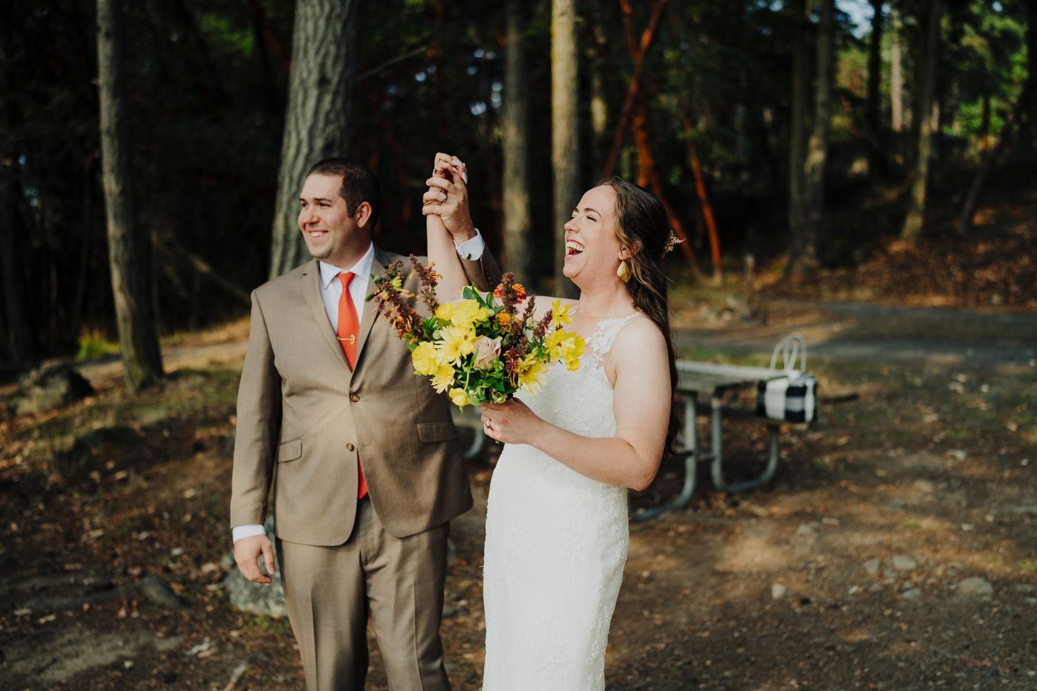 A joyous wedding couple shares a candid laugh while holding a vibrant yellow sunflower bouquet in a wooded setting.