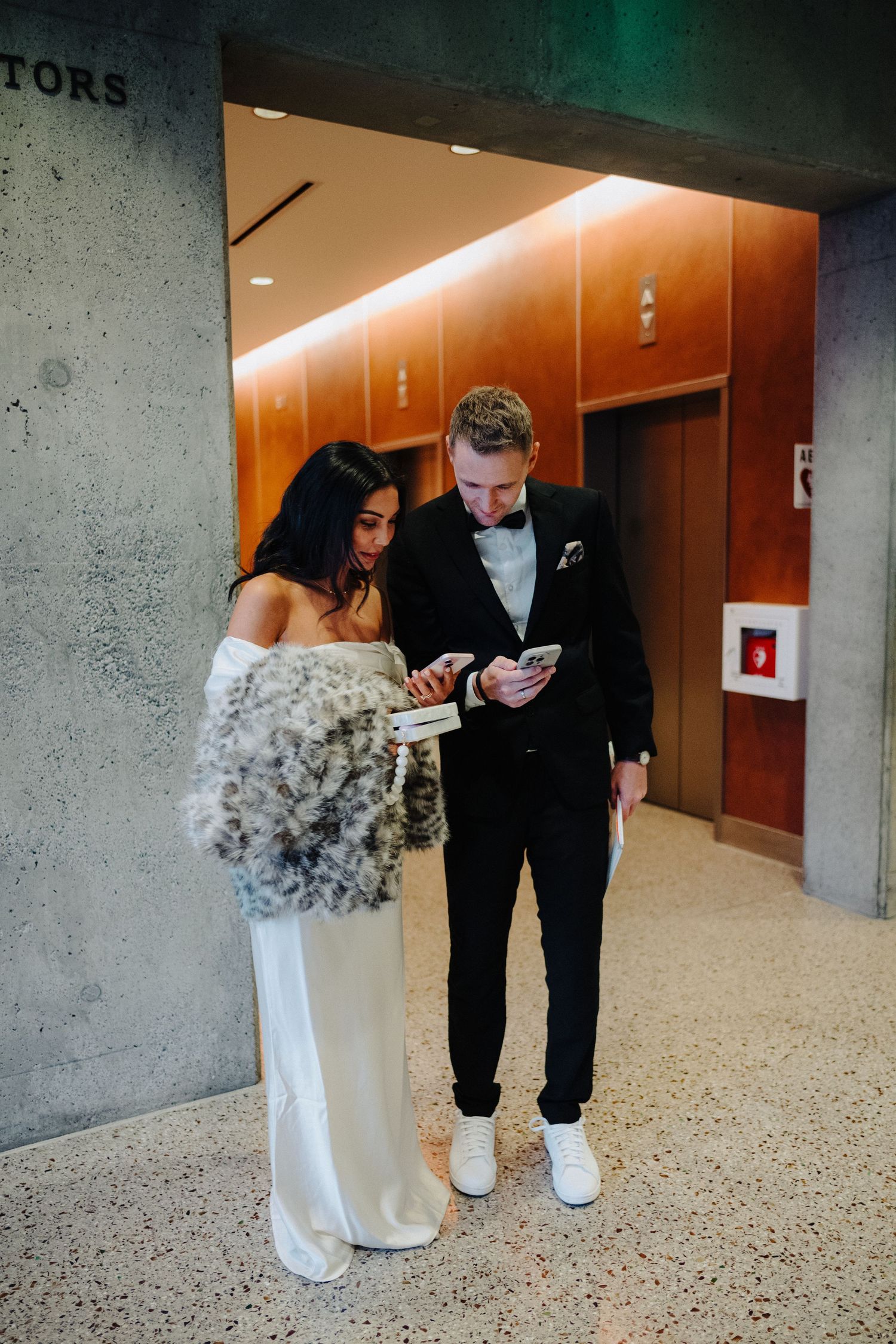 A couple checks their phones in a modern building hallway with warm lighting.