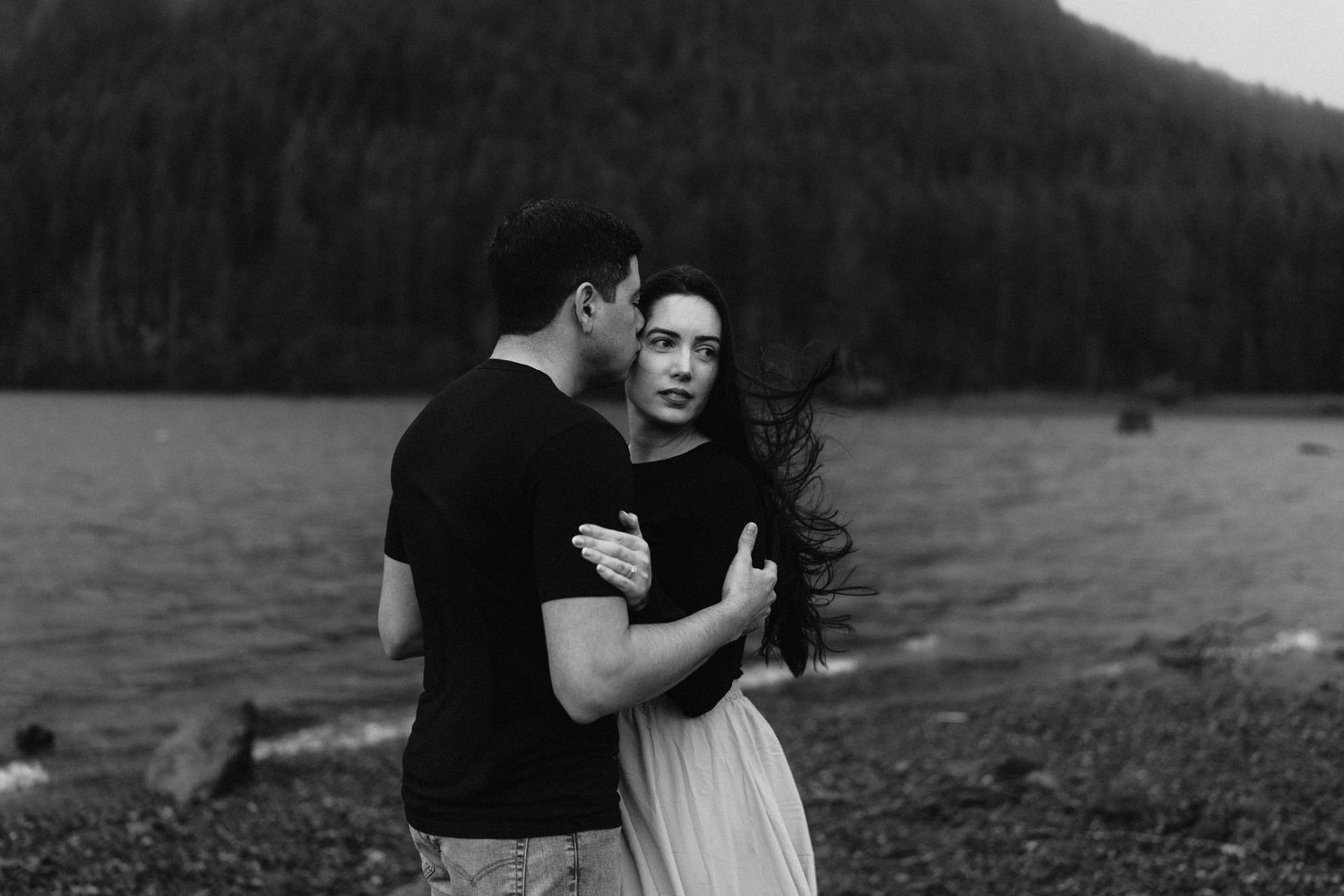 A tender black and white moment shared between a couple on a moody lakeside beach.