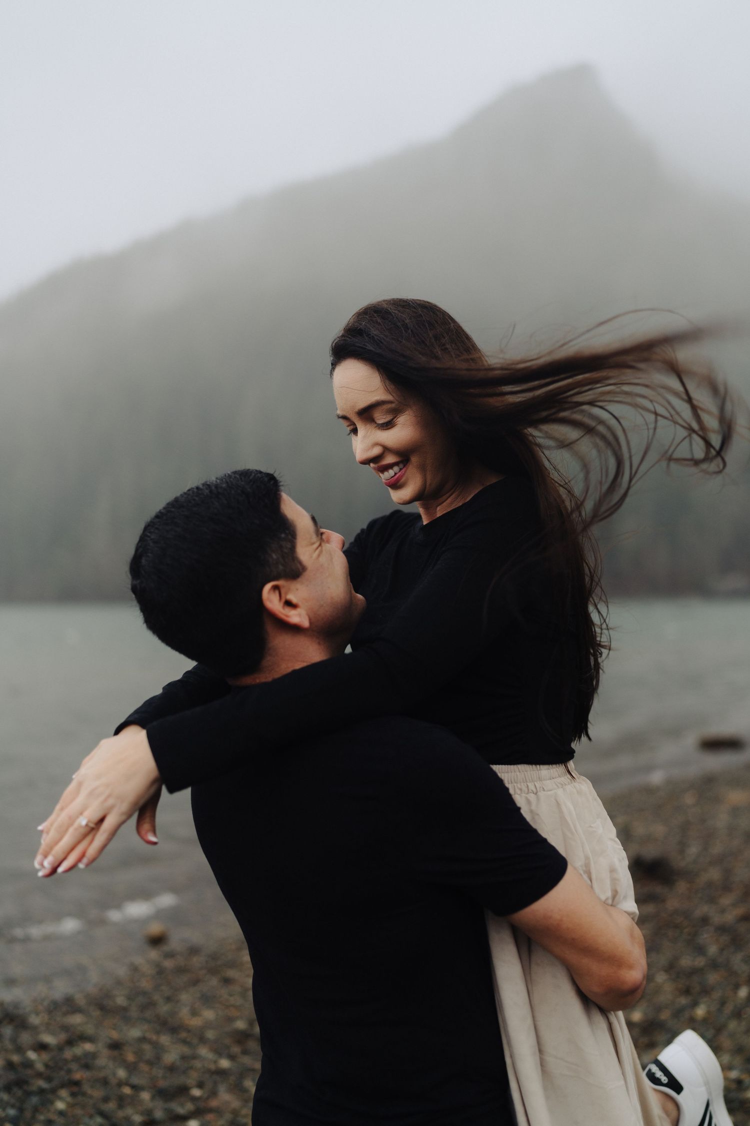 A romantic moment captured between a couple against a moody mountain backdrop on a rocky beach.