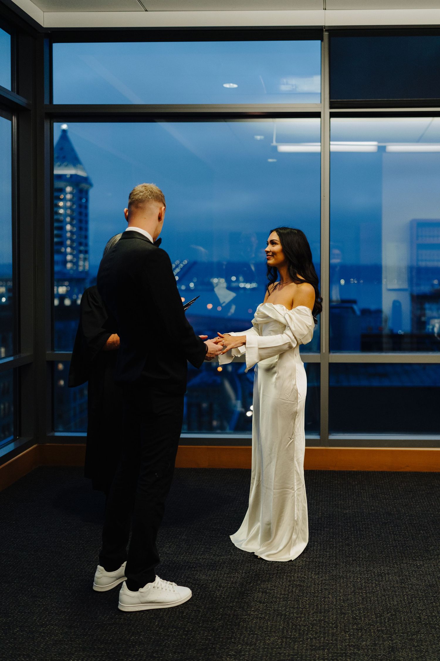 Bride in white gown and groom in black suit share an intimate moment against city skyline backdrop.