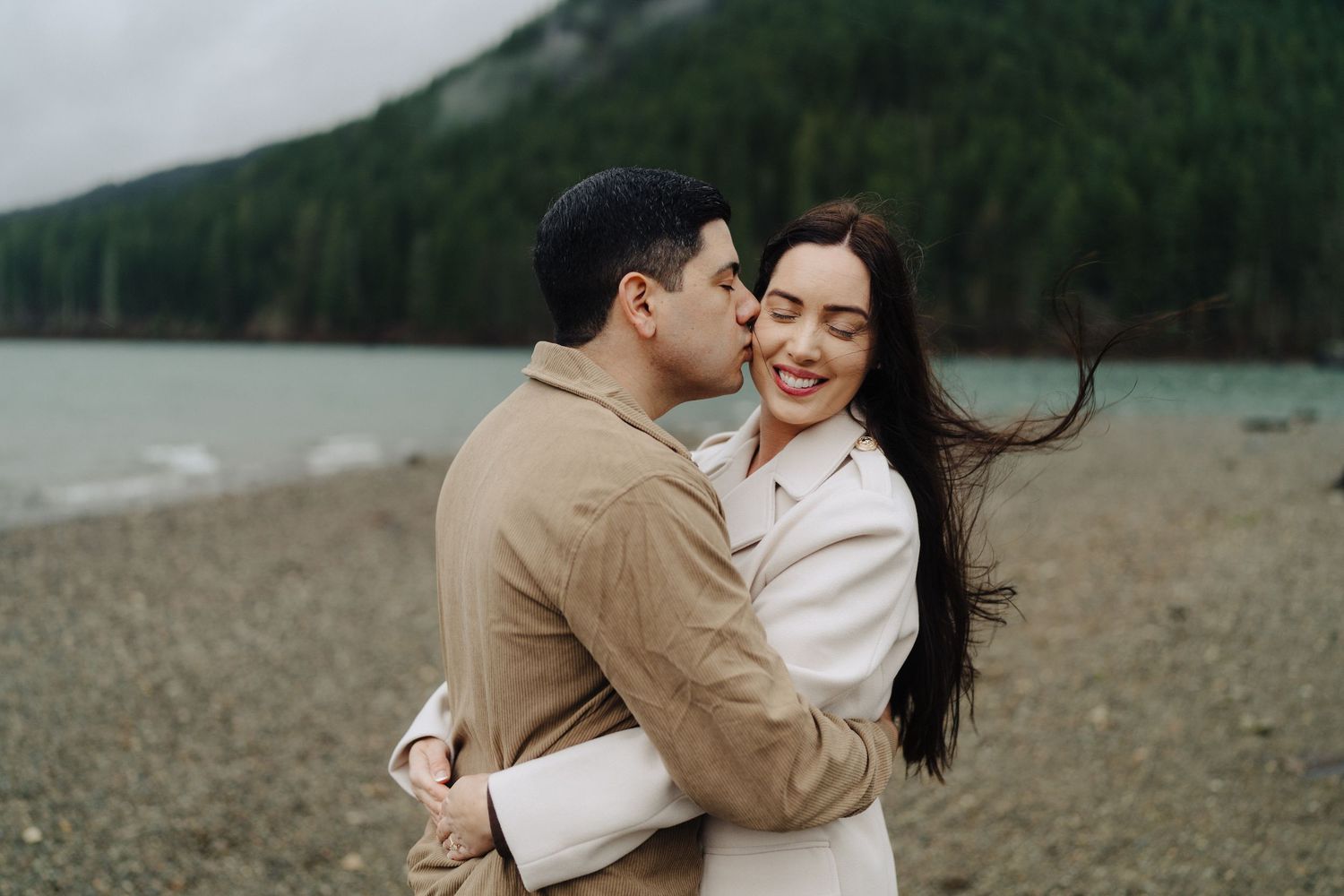 Joyful couple laughs while embracing on rocky beach with misty mountain scenery.