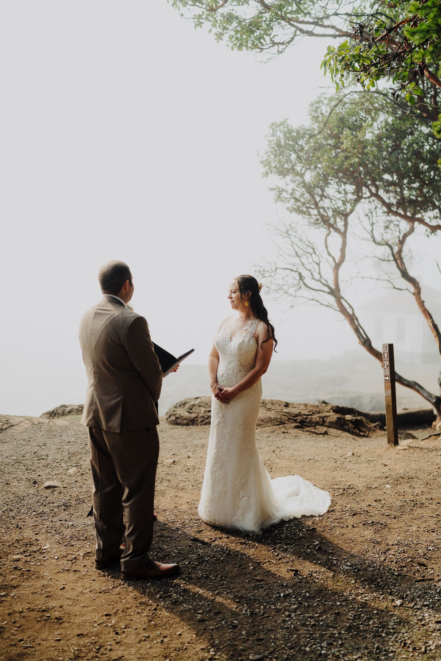 A wedding ceremony unfolds on a foggy hilltop with sparse trees in the background.