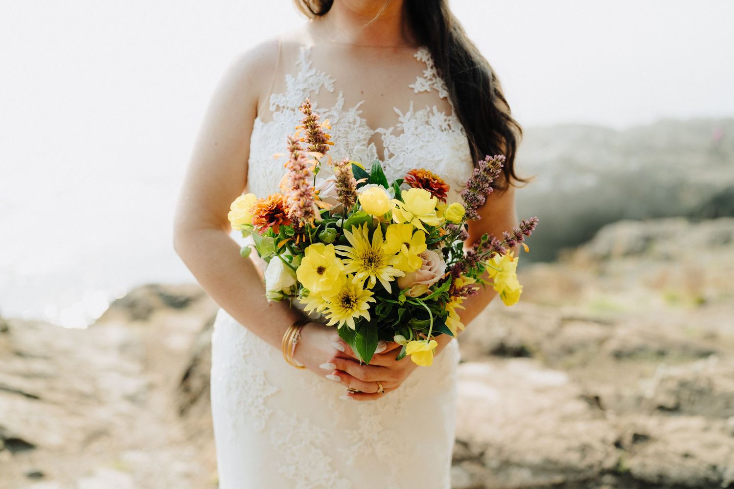 Bright yellow sunflowers and wildflowers in a bridal bouquet against a lace dress.