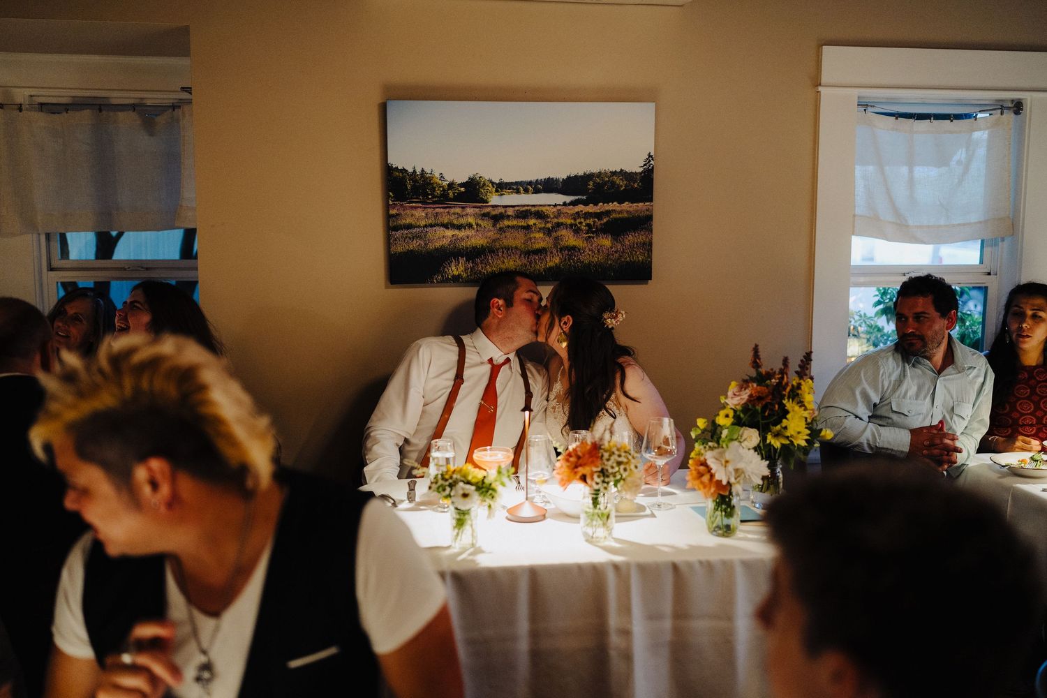 A group dines at a rustic reception with landscape artwork and warm lighting in the background.