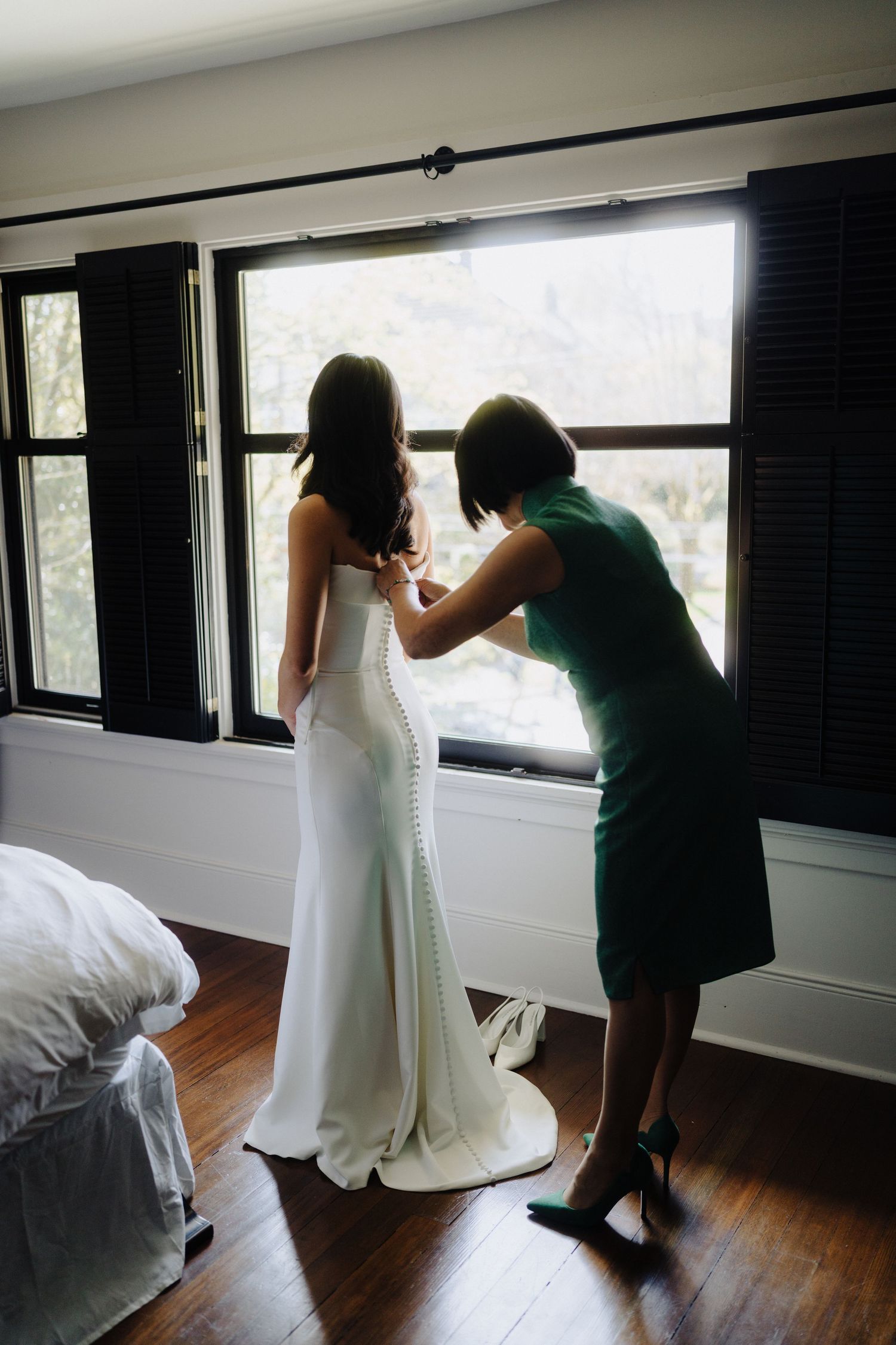 A white wedding dress being adjusted near a bright window in a room with hardwood floors.