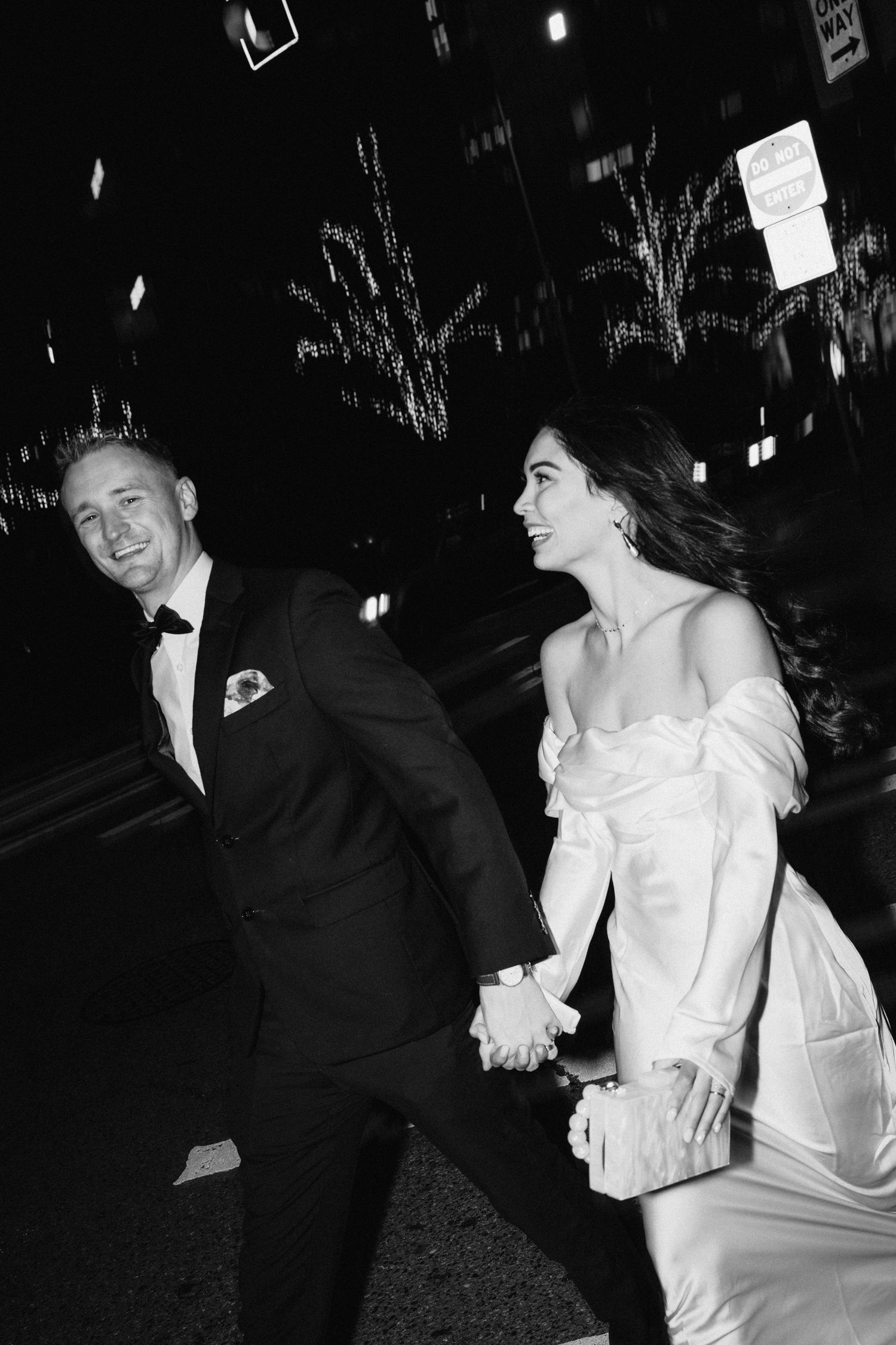 Black and white photo of newlyweds running joyfully through city streets at night.