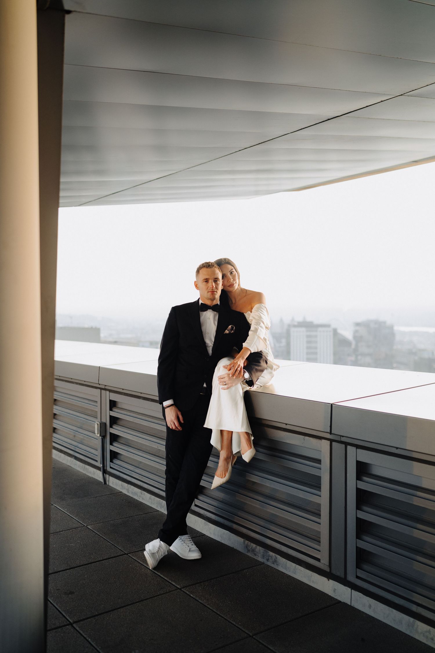 A couple poses against a railing on a modern building terrace with city views in the background.