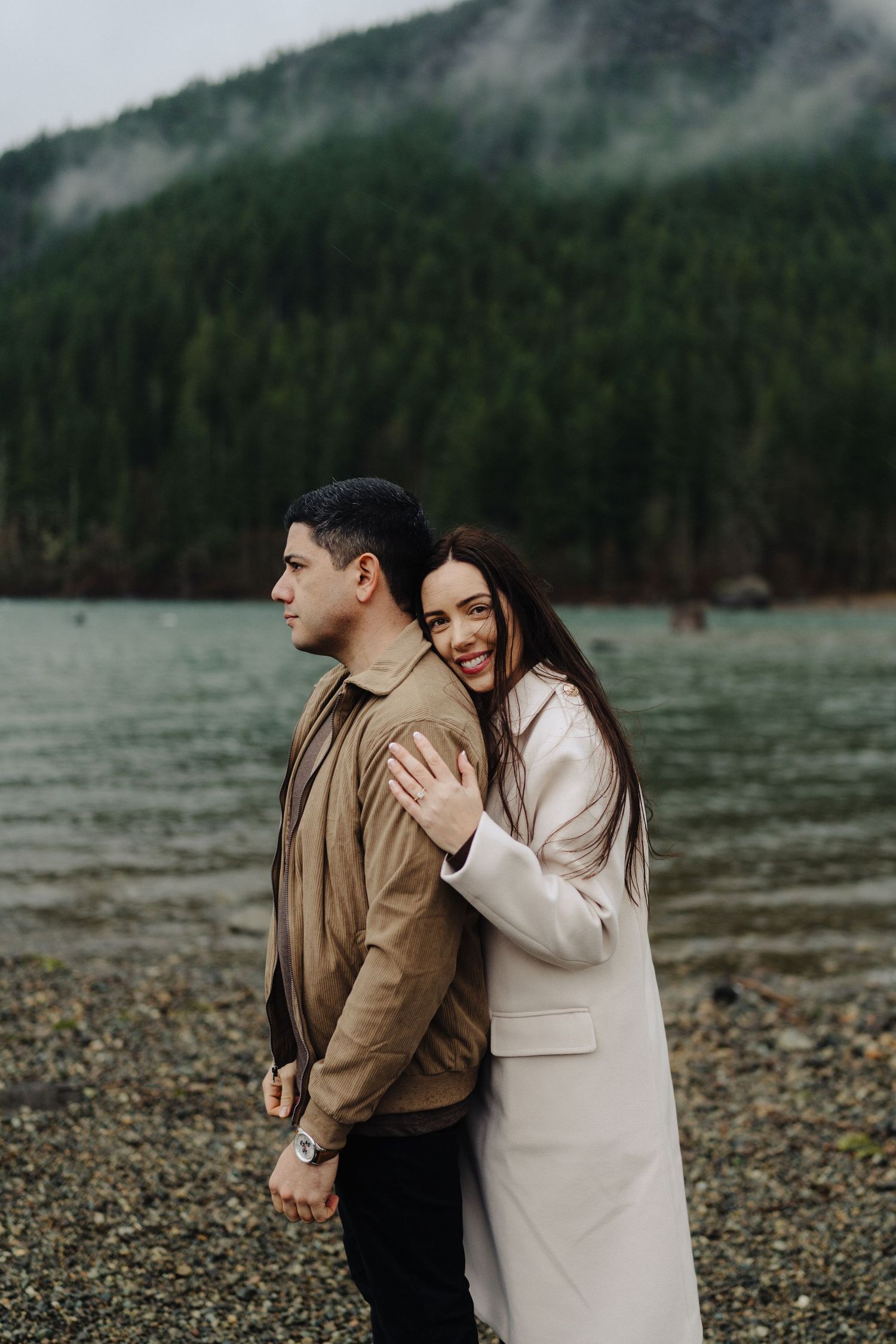 Two people share a tender moment on a pebble beach with foggy mountains and lake in the background.