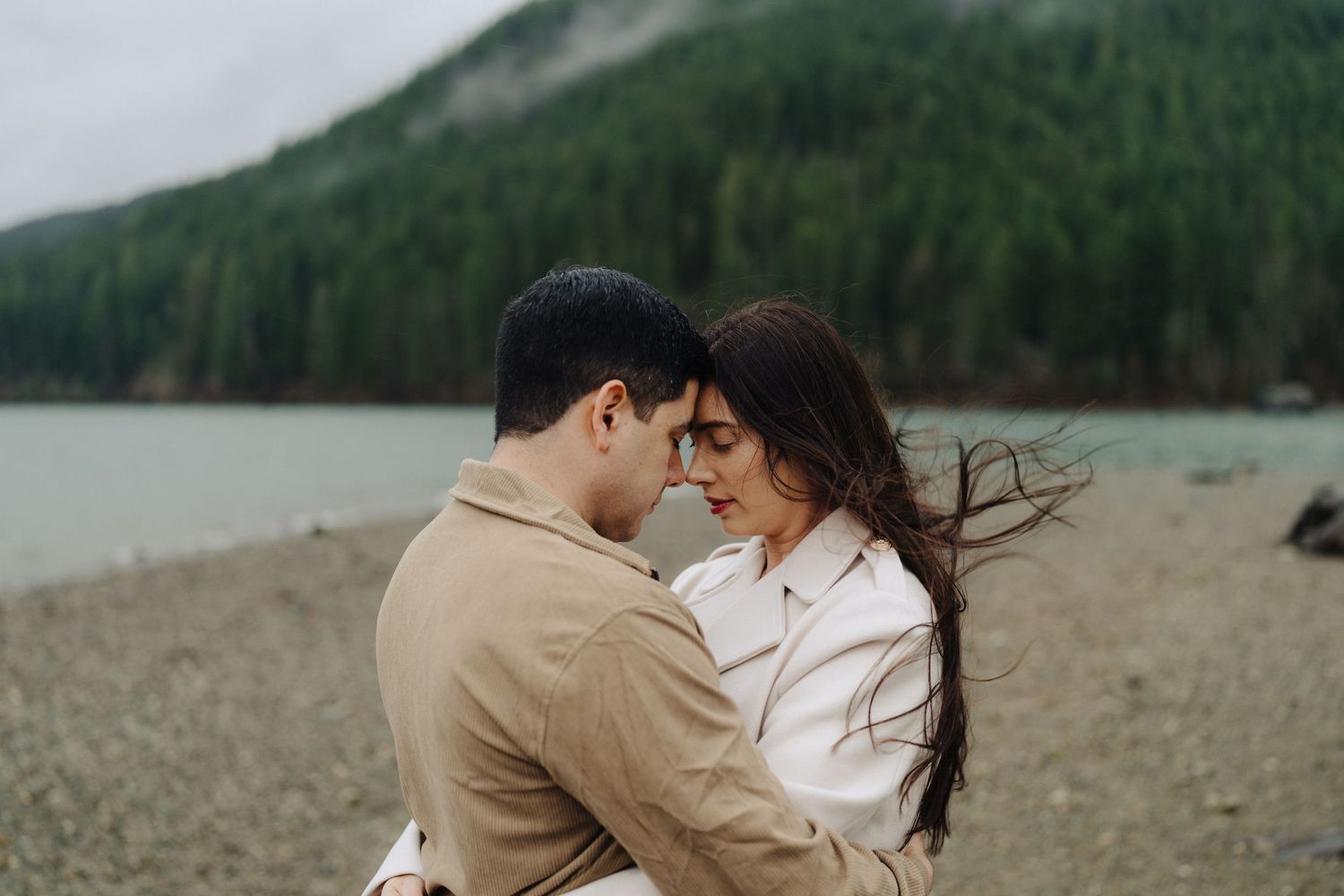 A romantic couple embraces on a rocky beach with forested mountains and cloudy skies in the background.