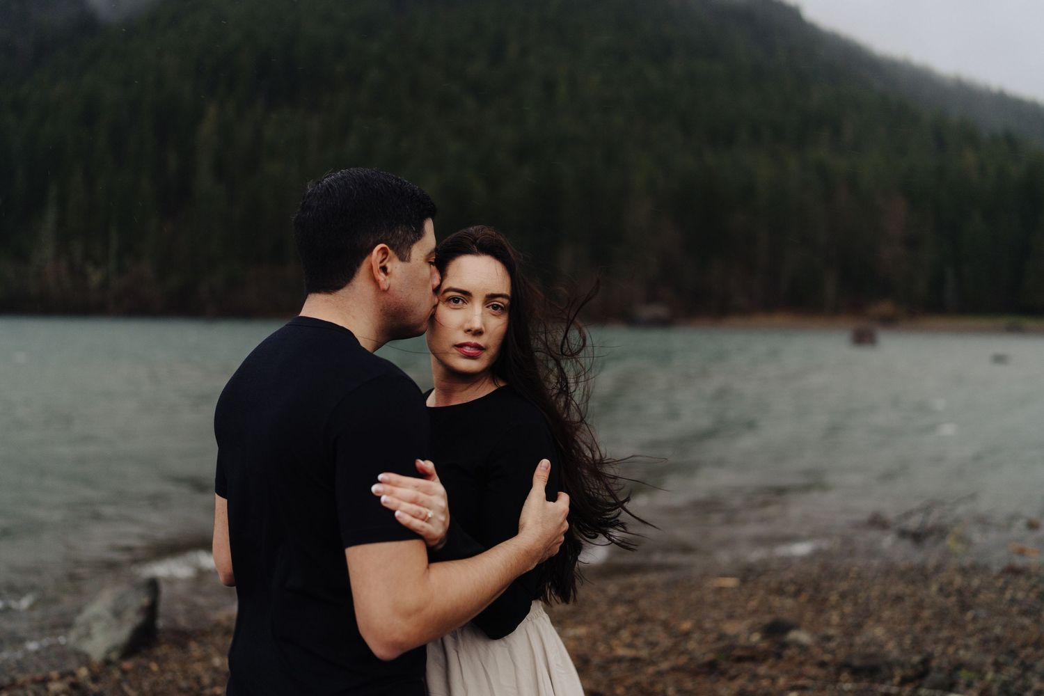 Intimate couple sharing tender moment on moody lakeside with mountains in background.