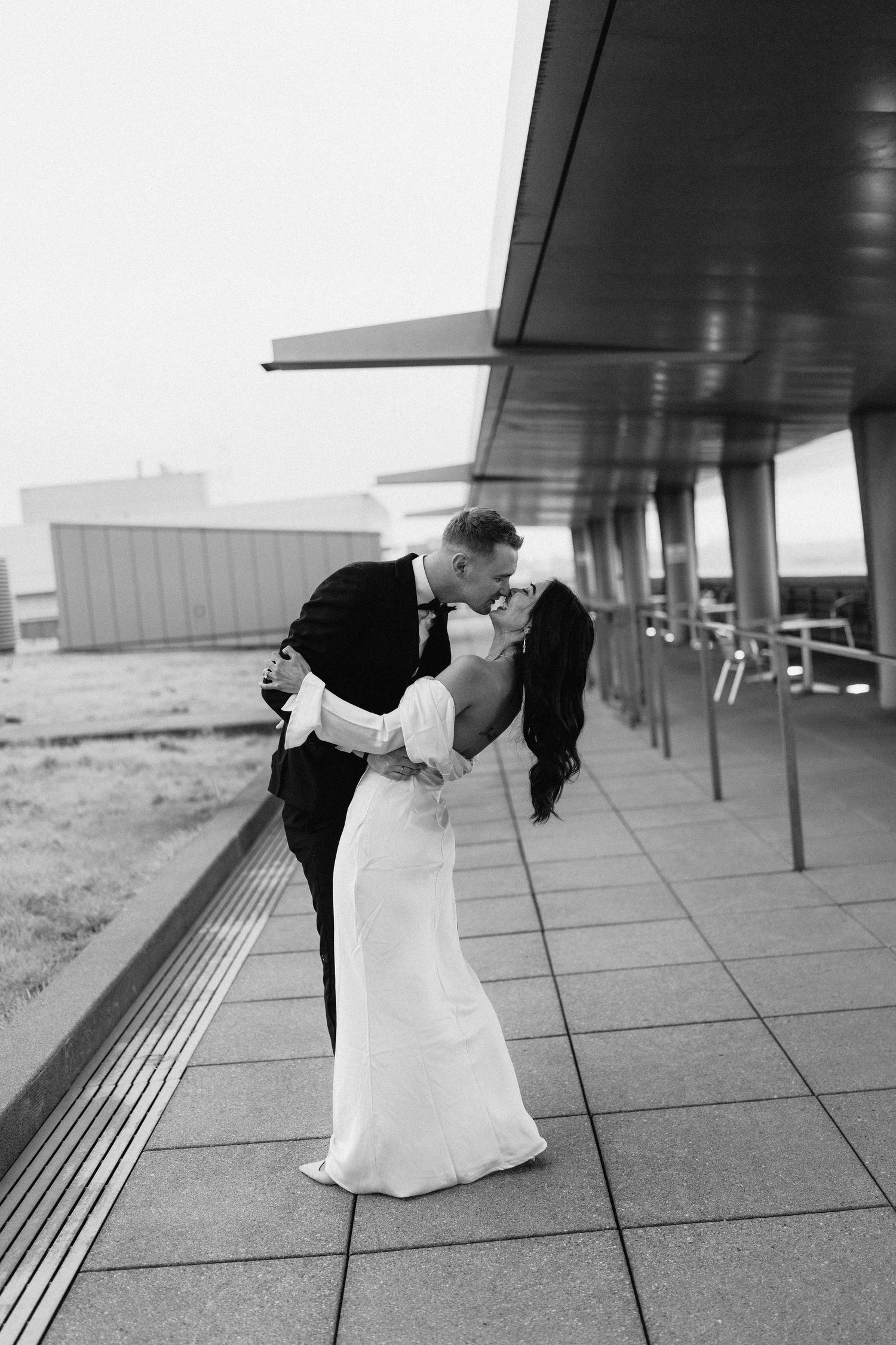 A couple in formal wear shares a romantic dip kiss on an outdoor terrace in black and white.