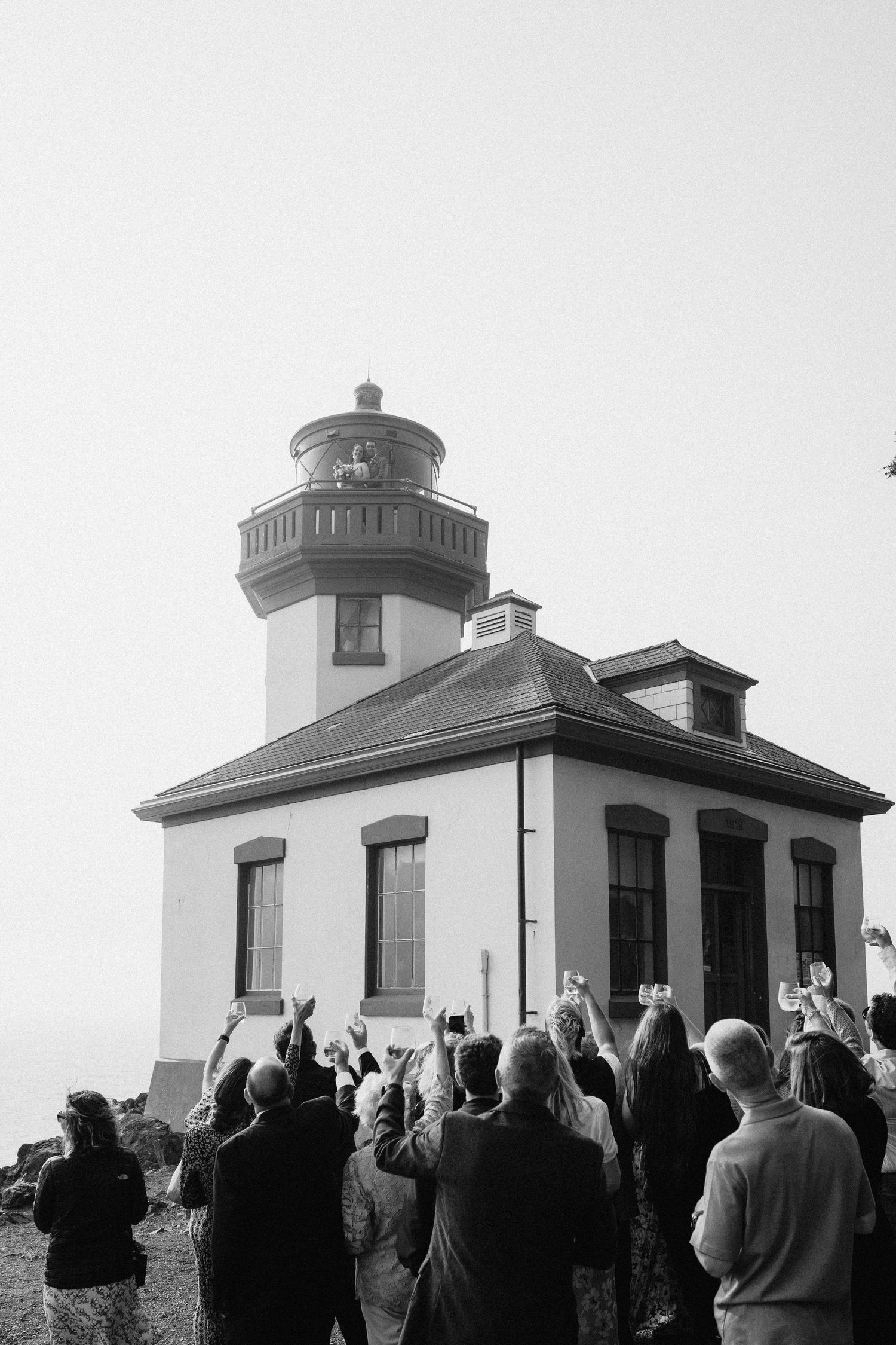 Wedding guests gather outside a historic white lighthouse for an outdoor ceremony.