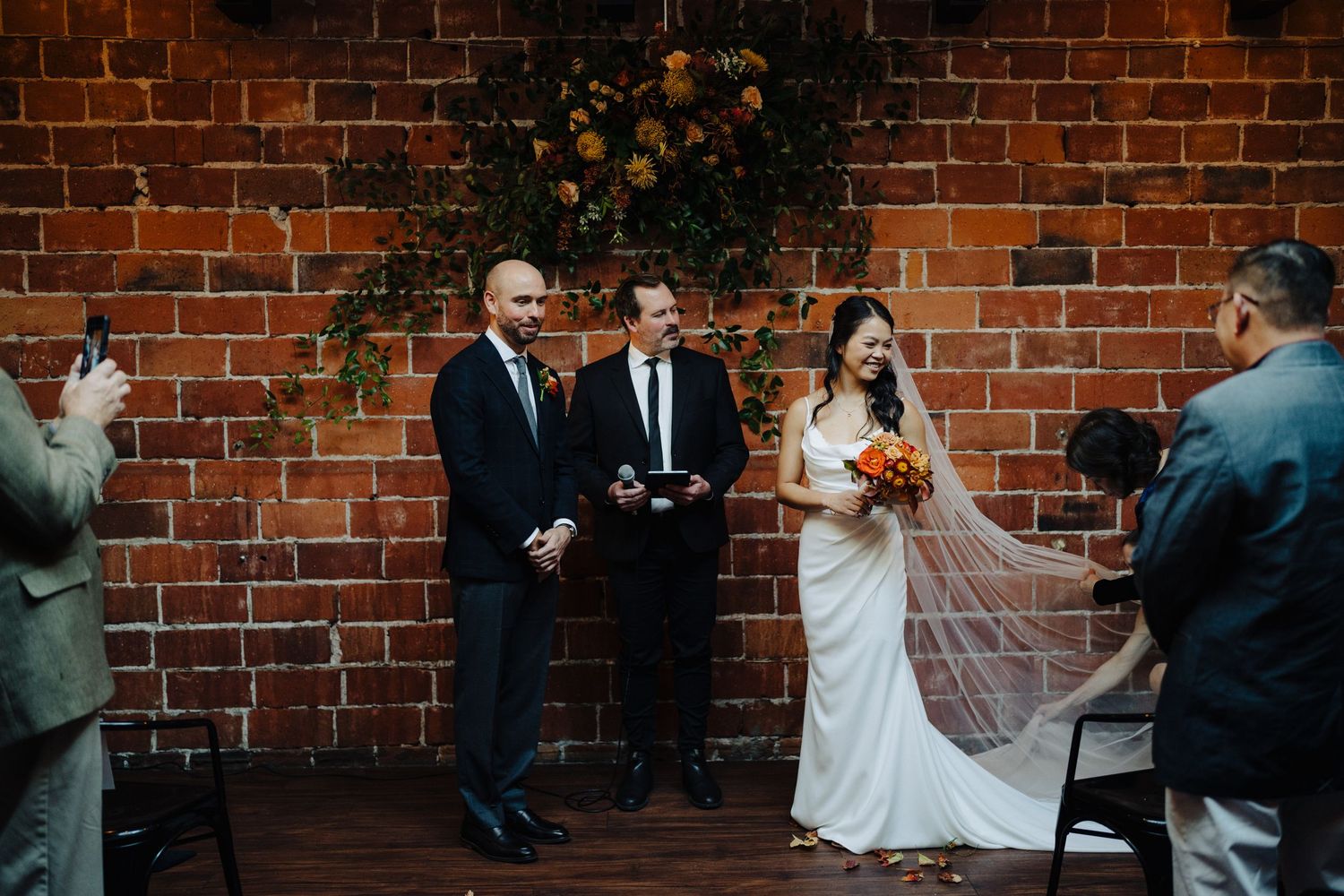 Wedding ceremony in front of rustic brick wall decorated with floral arrangements.