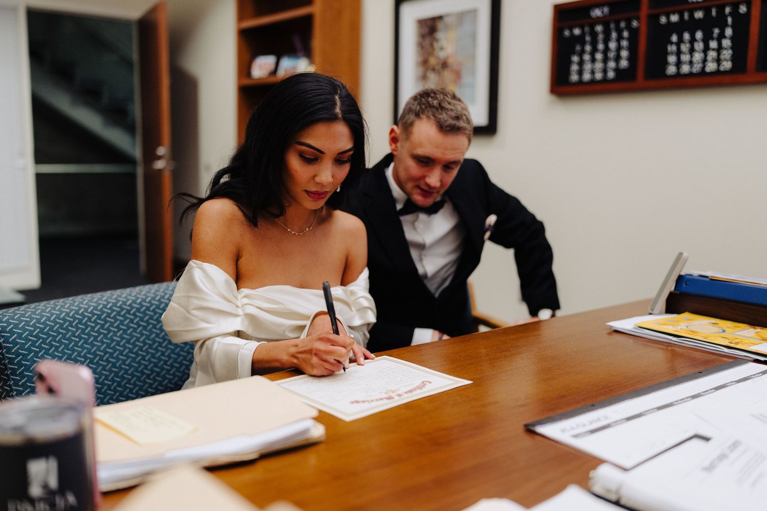 A couple reviewing and signing documents together at a wooden desk in an office setting.