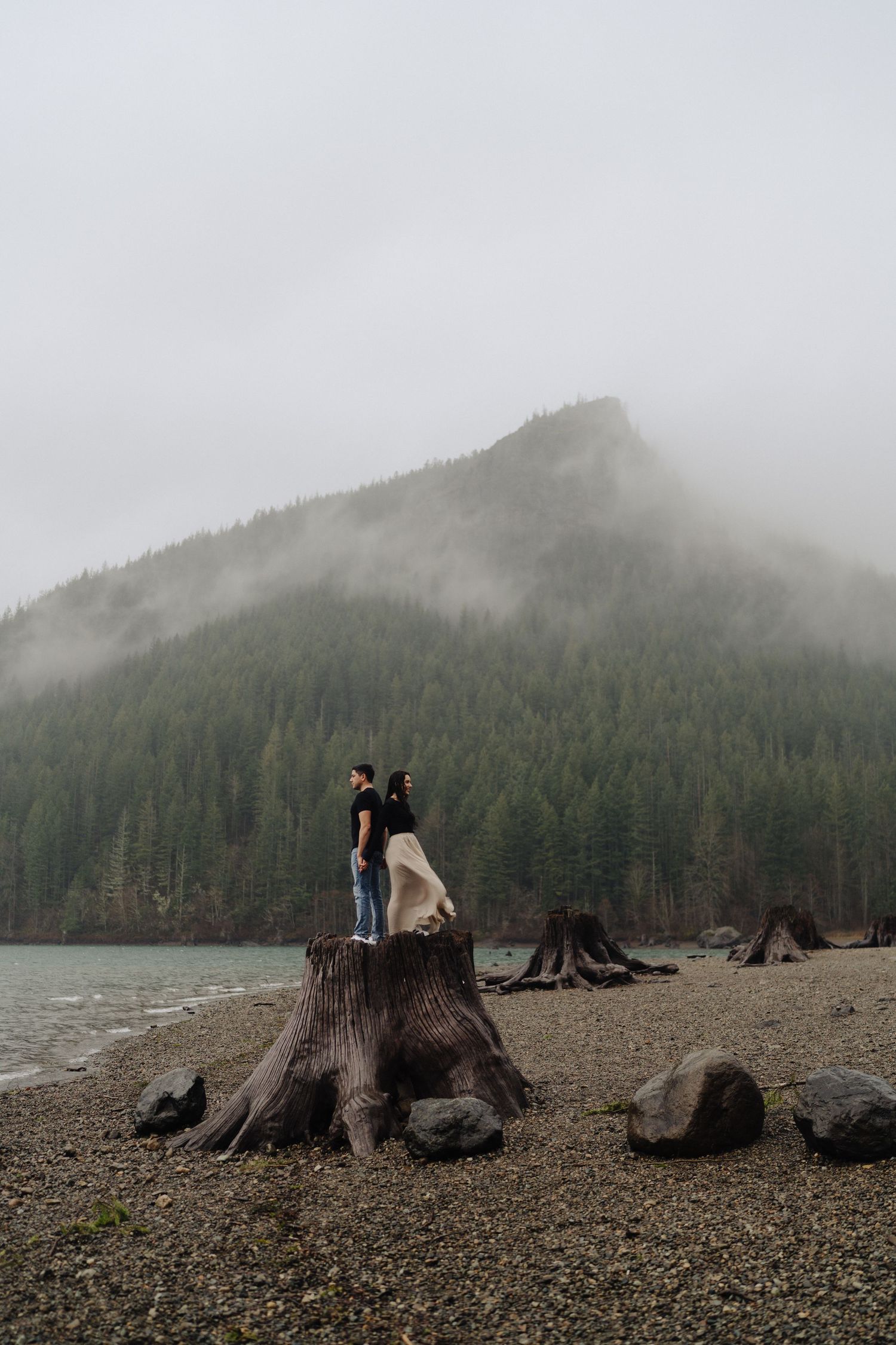 Two people pose together atop a giant tree stump with foggy mountains and lake in the distance.