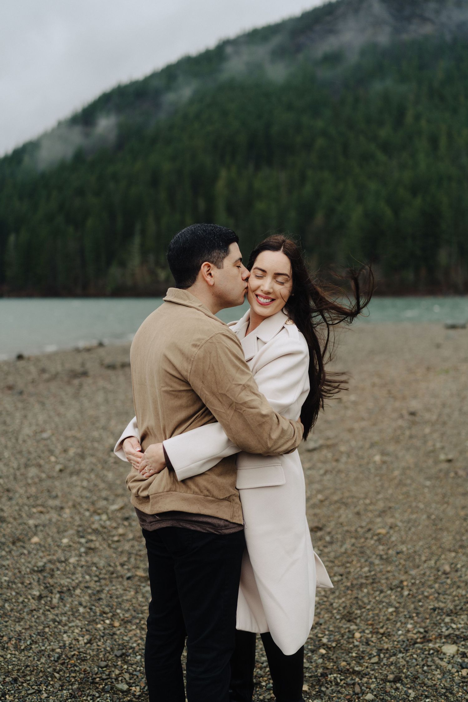 Happy couple embraces on pebble beach with forest mountain backdrop on overcast day.