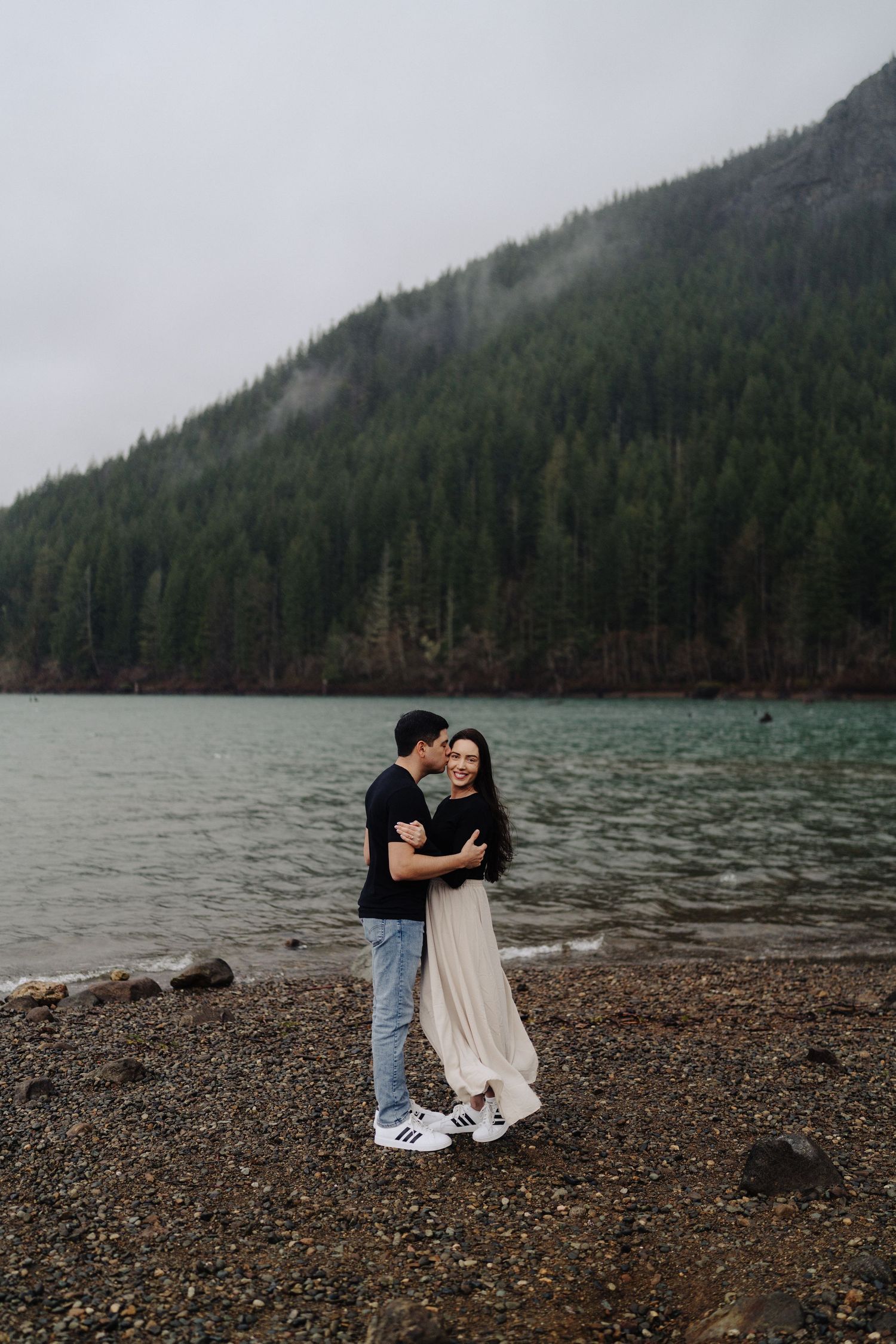 A couple embraces on a rocky beach shore with misty evergreen mountains and a lake in the background.