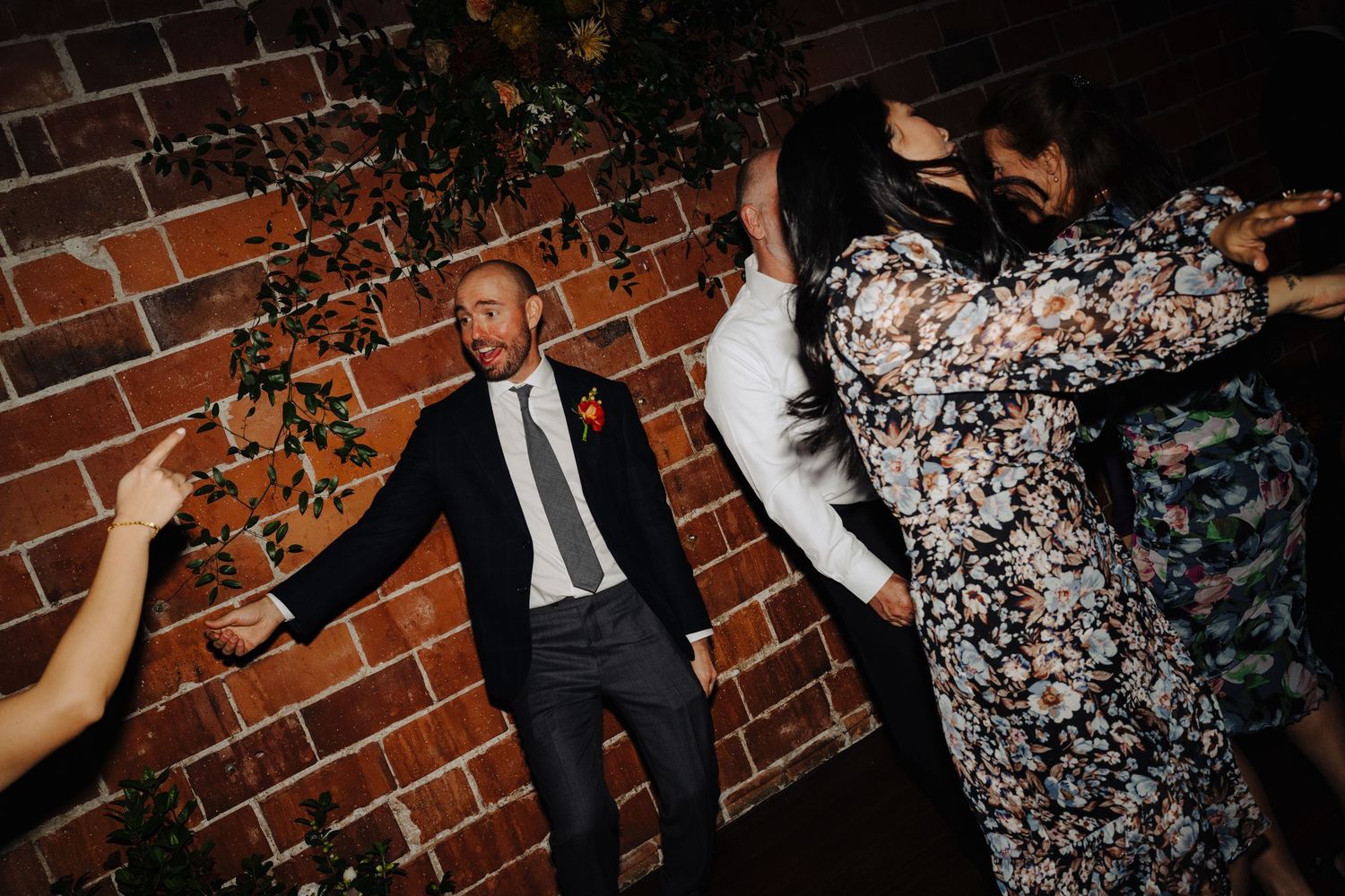 Wedding party dancing energetically against exposed brick wall with decorative greenery.