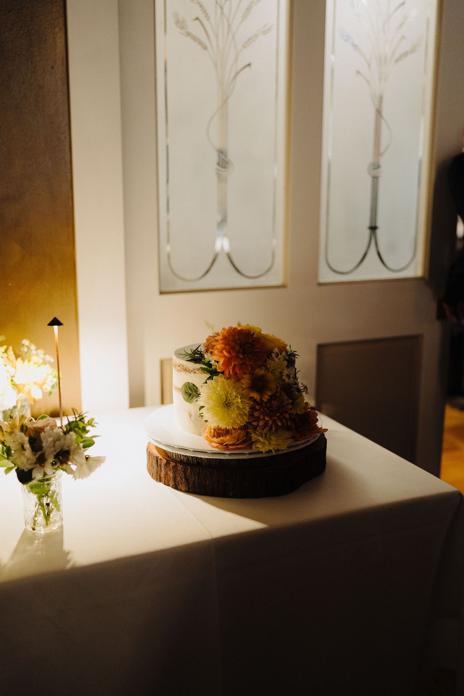 A rustic wedding cake with orange and yellow flowers sits on a wooden slice in an elegant white room.