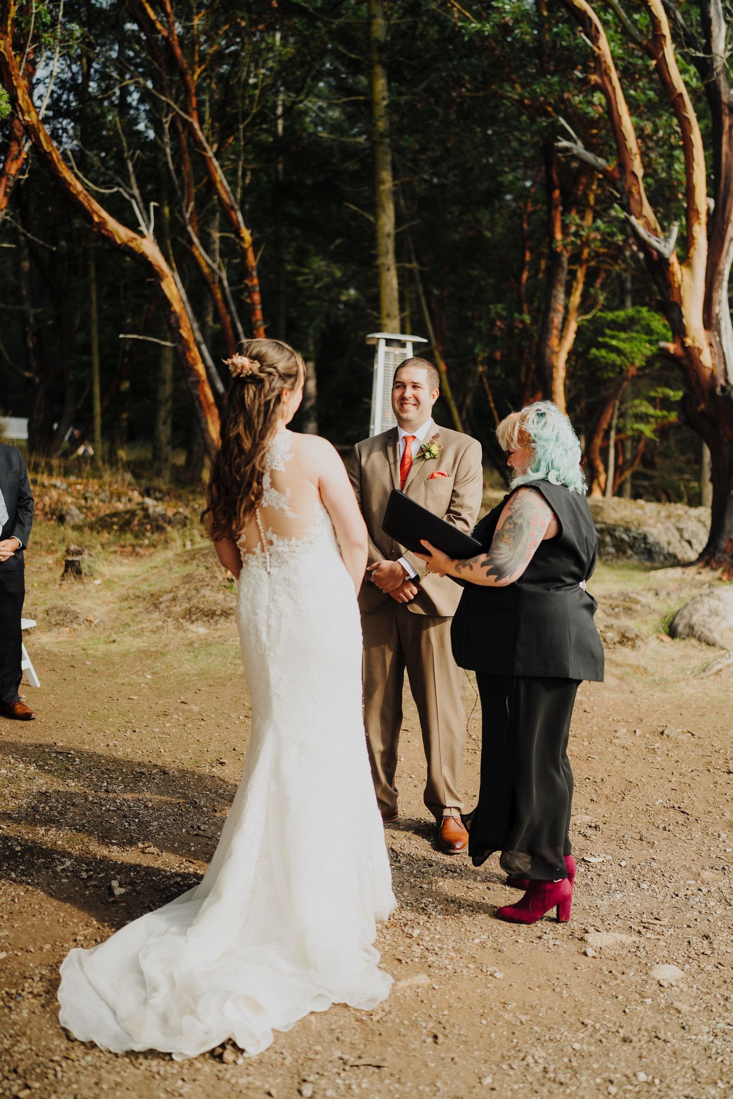 An intimate outdoor wedding ceremony takes place in a wooded setting with guests standing in a circle.