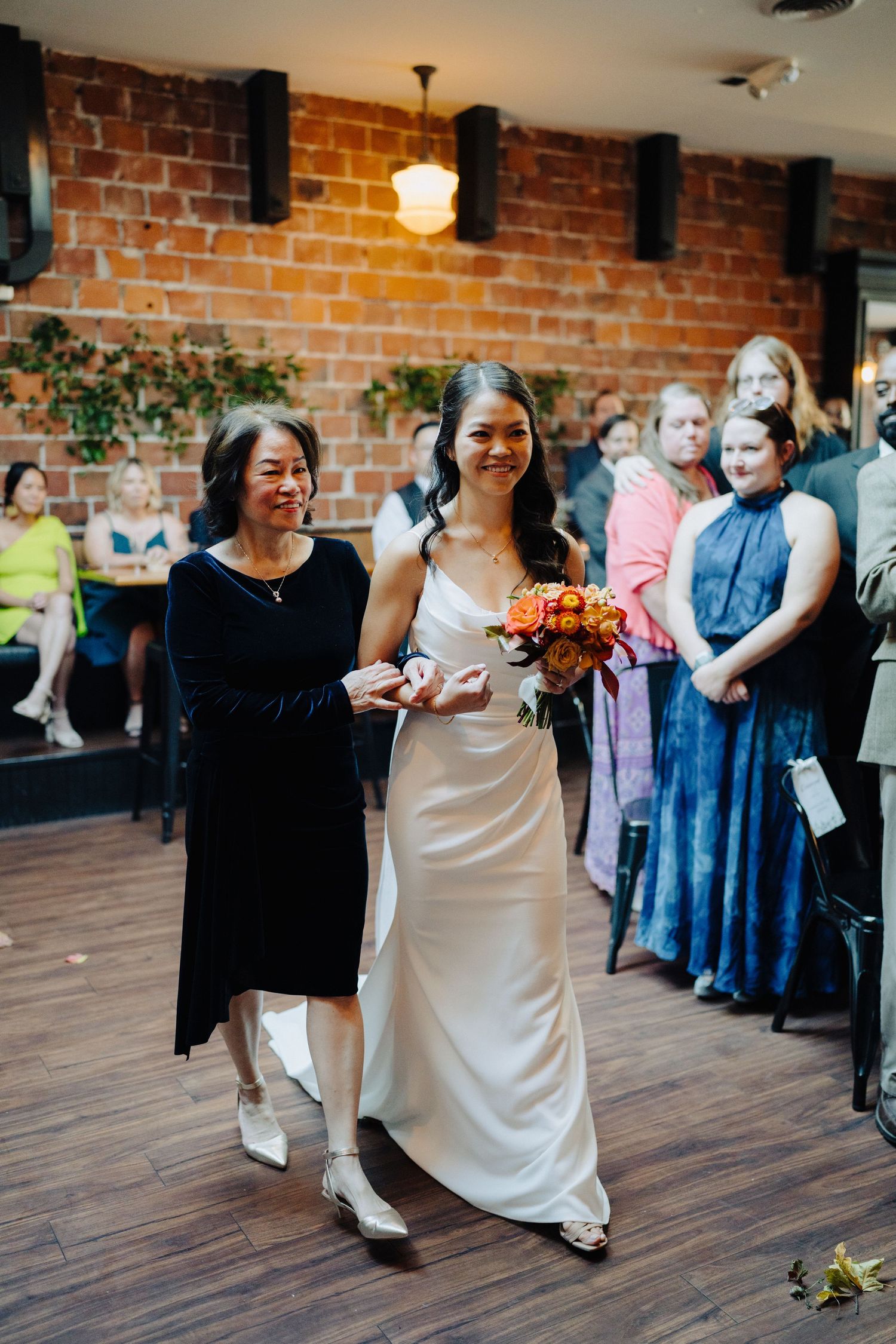 A wedding ceremony in a rustic brick-walled venue with guests watching the processional.