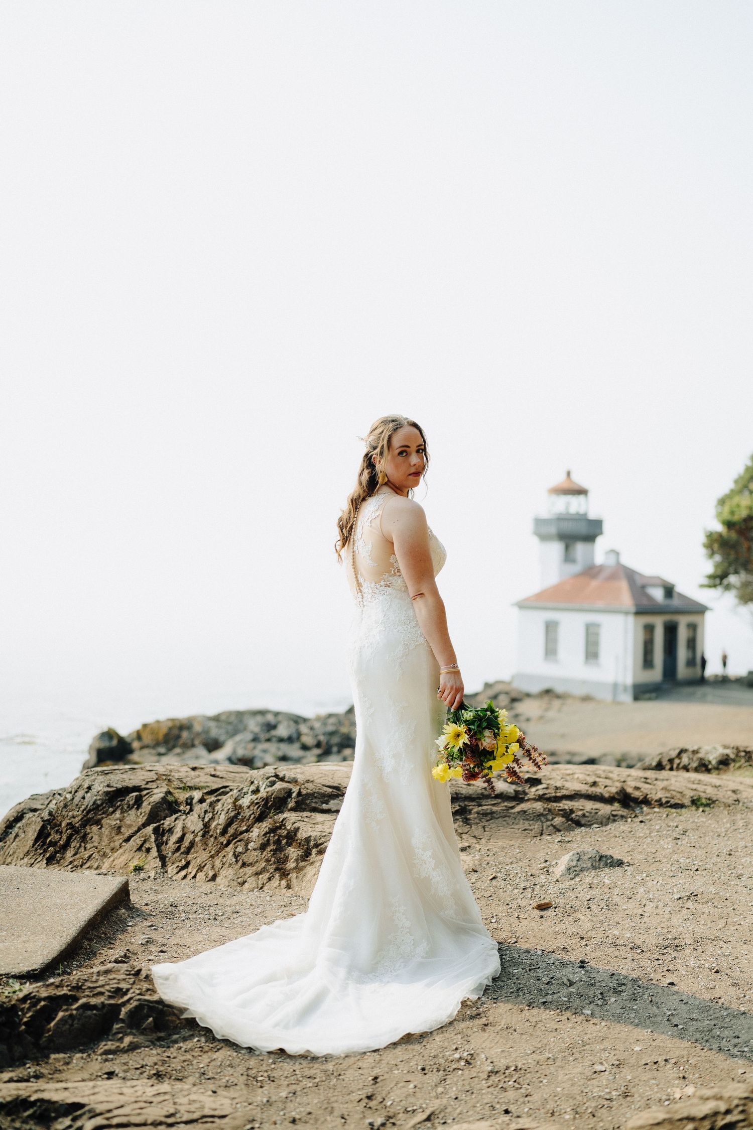 White lighthouse stands atop rocky cliffs near the ocean with wedding dress in foreground.