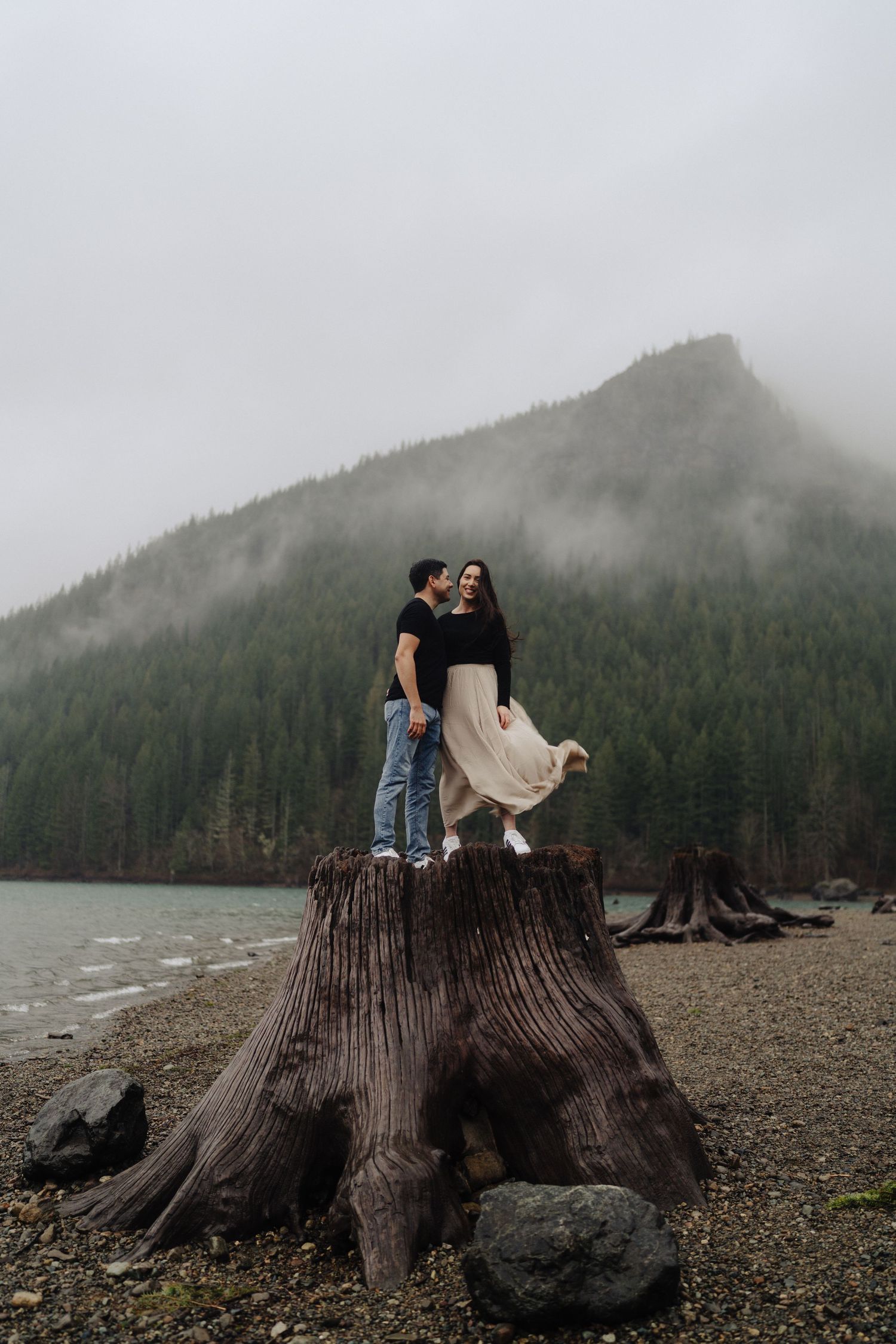 A couple stands on a large tree stump by a misty mountain lake with evergreen forests in the background.
