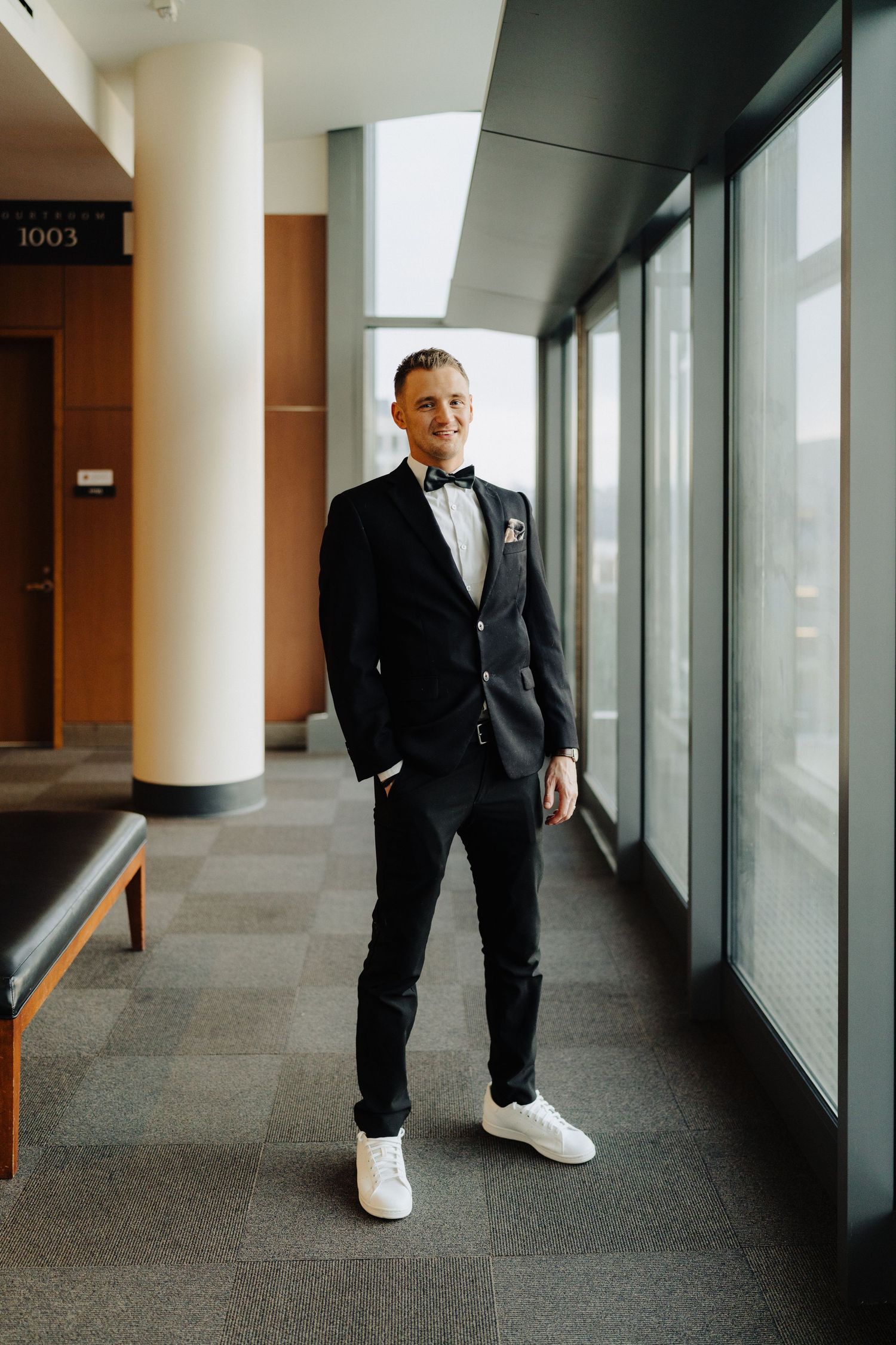 Groom in black tuxedo and white sneakers poses confidently in modern hallway.