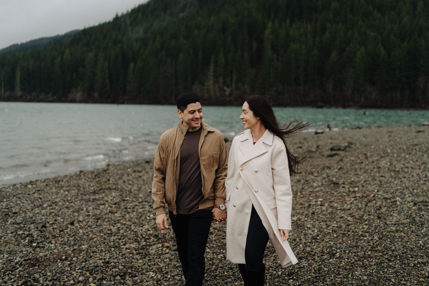 A couple walks along a rocky lakeshore with evergreen-covered mountains rising in the background.
