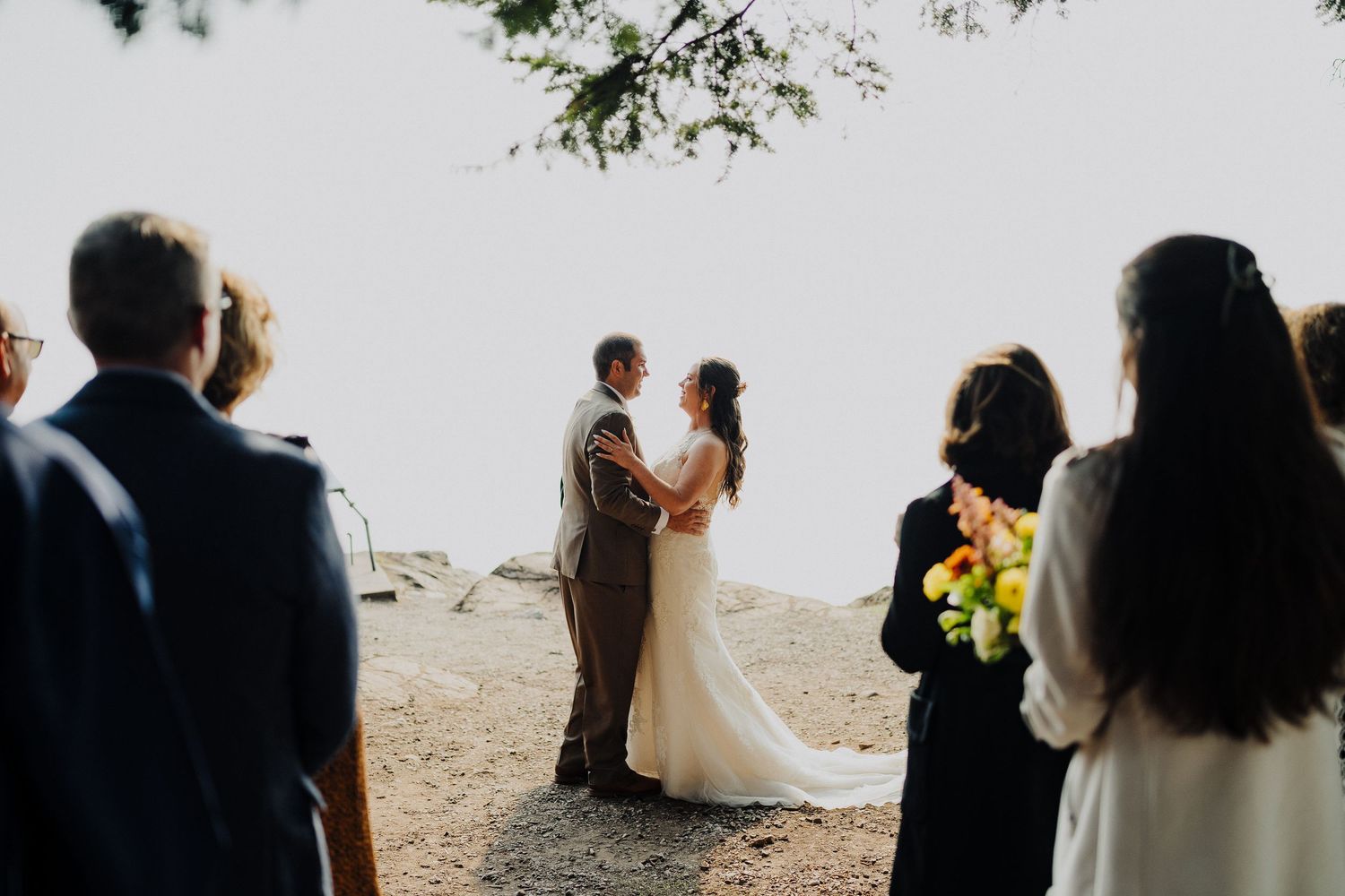 Wedding ceremony taking place on cliff overlooking ocean with guests watching couple during sunset ceremony.