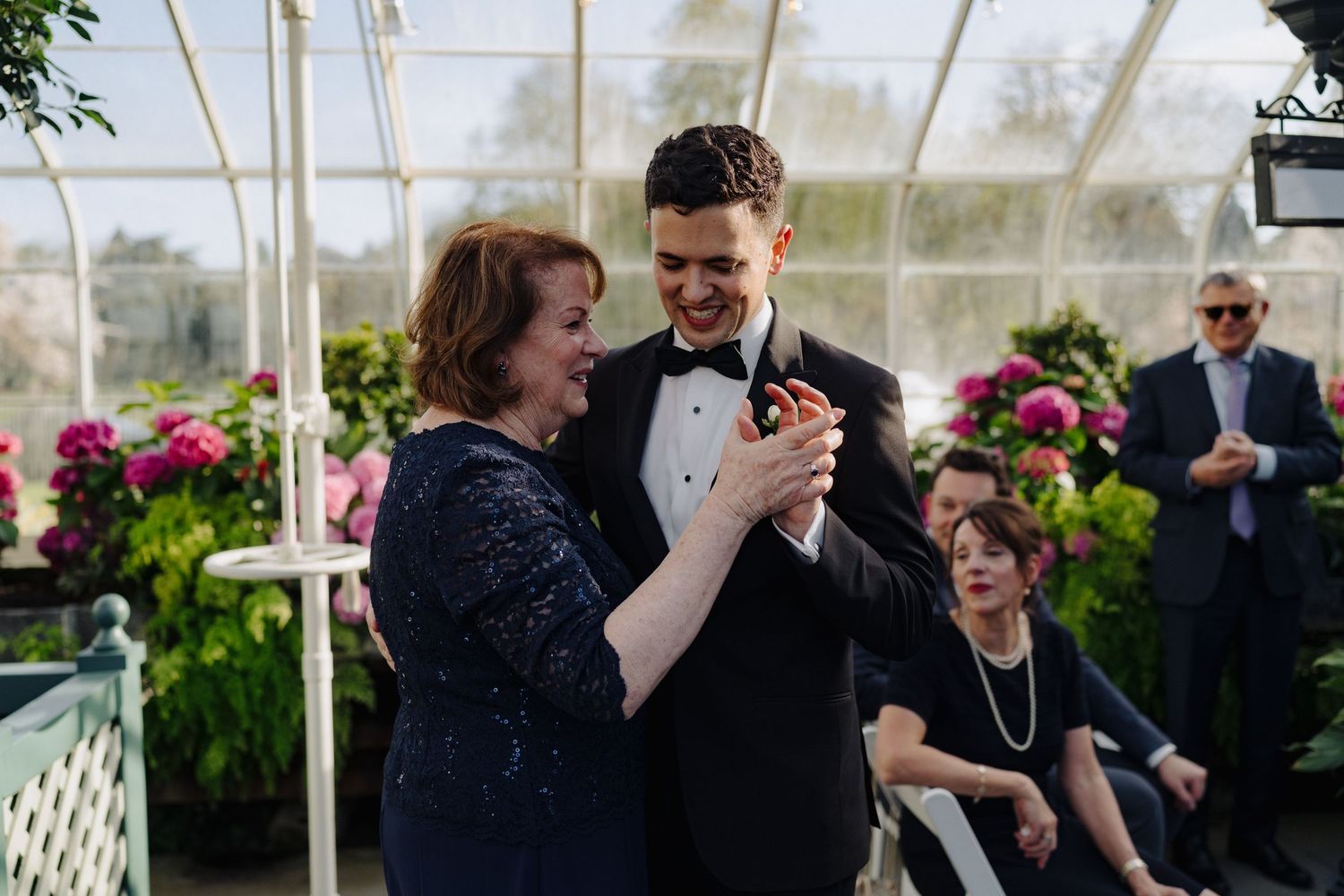 Wedding guests dancing together in a decorated greenhouse venue with pink flowers and natural light.