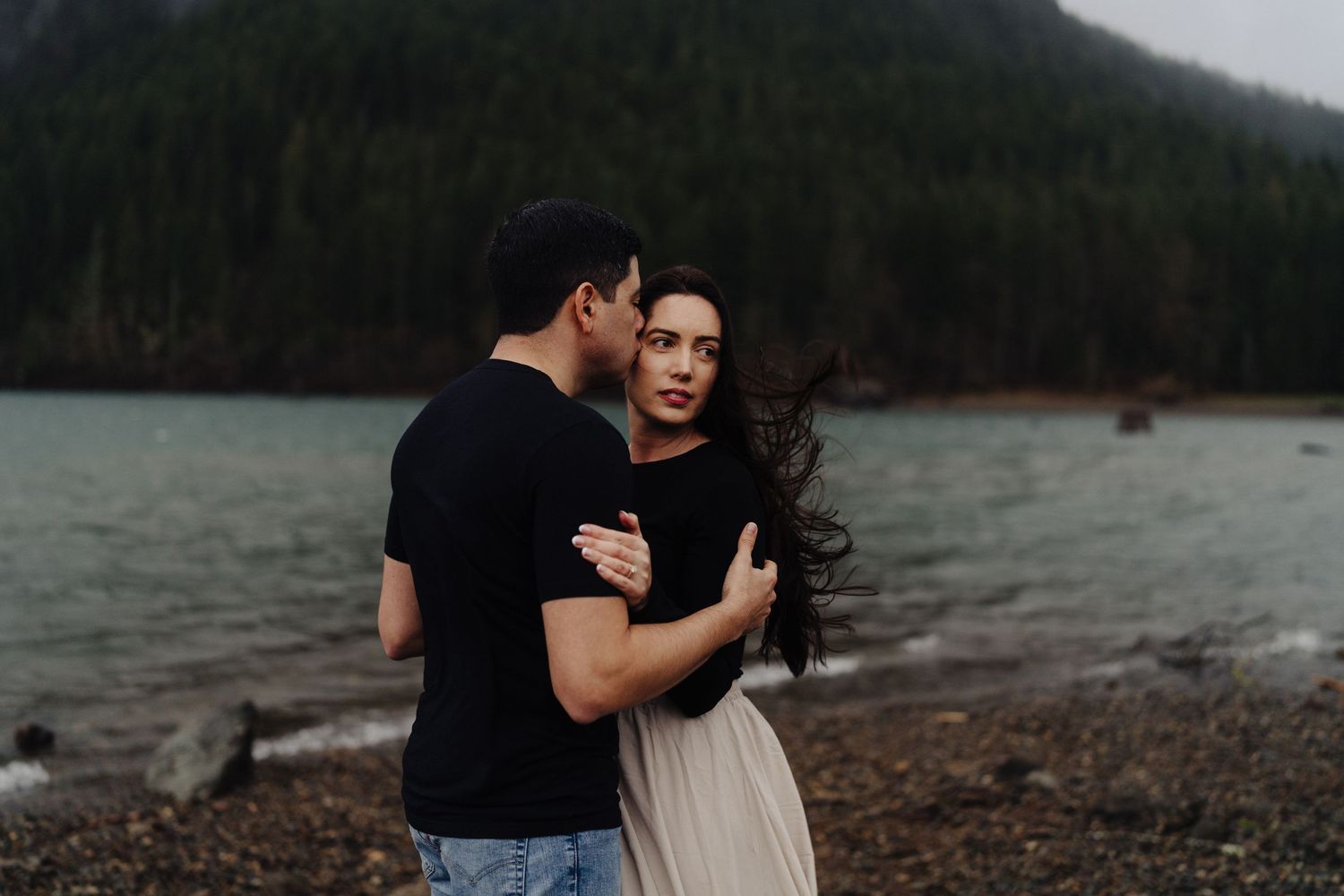 A couple embraces on a rocky shoreline near a misty lake surrounded by evergreen-covered mountains.