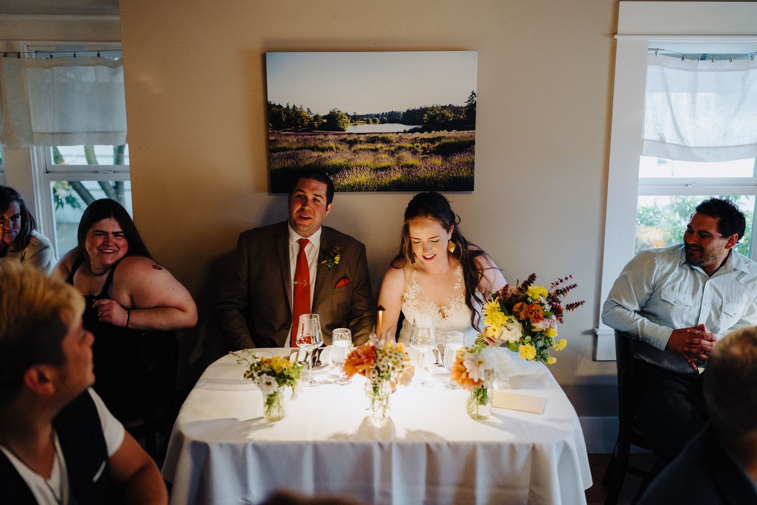 People gathered at a formal event around a dining table with flowers and decor in a bright room with landscape art.