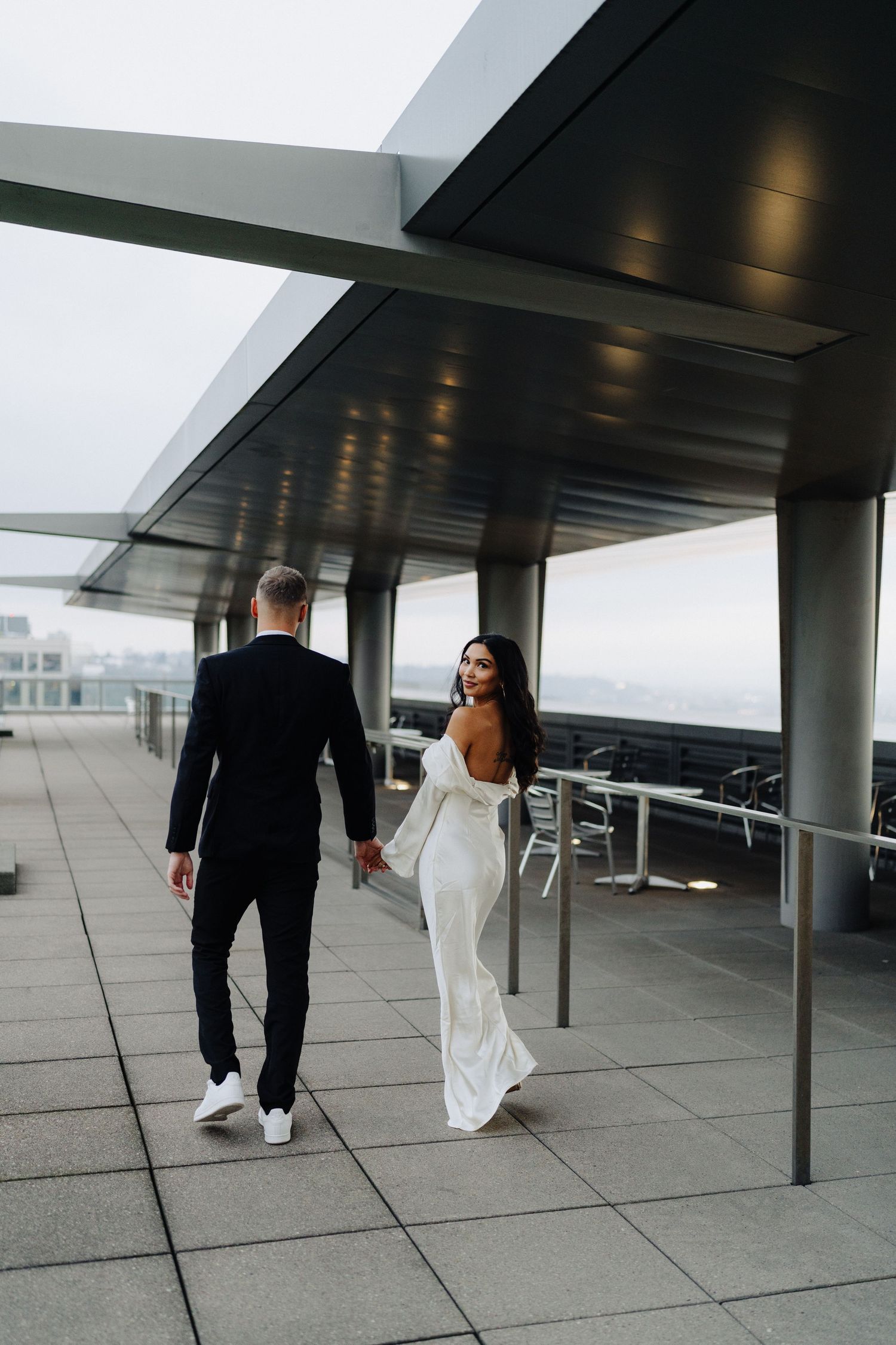 A couple in wedding attire walks hand in hand on a modern rooftop terrace with city views.