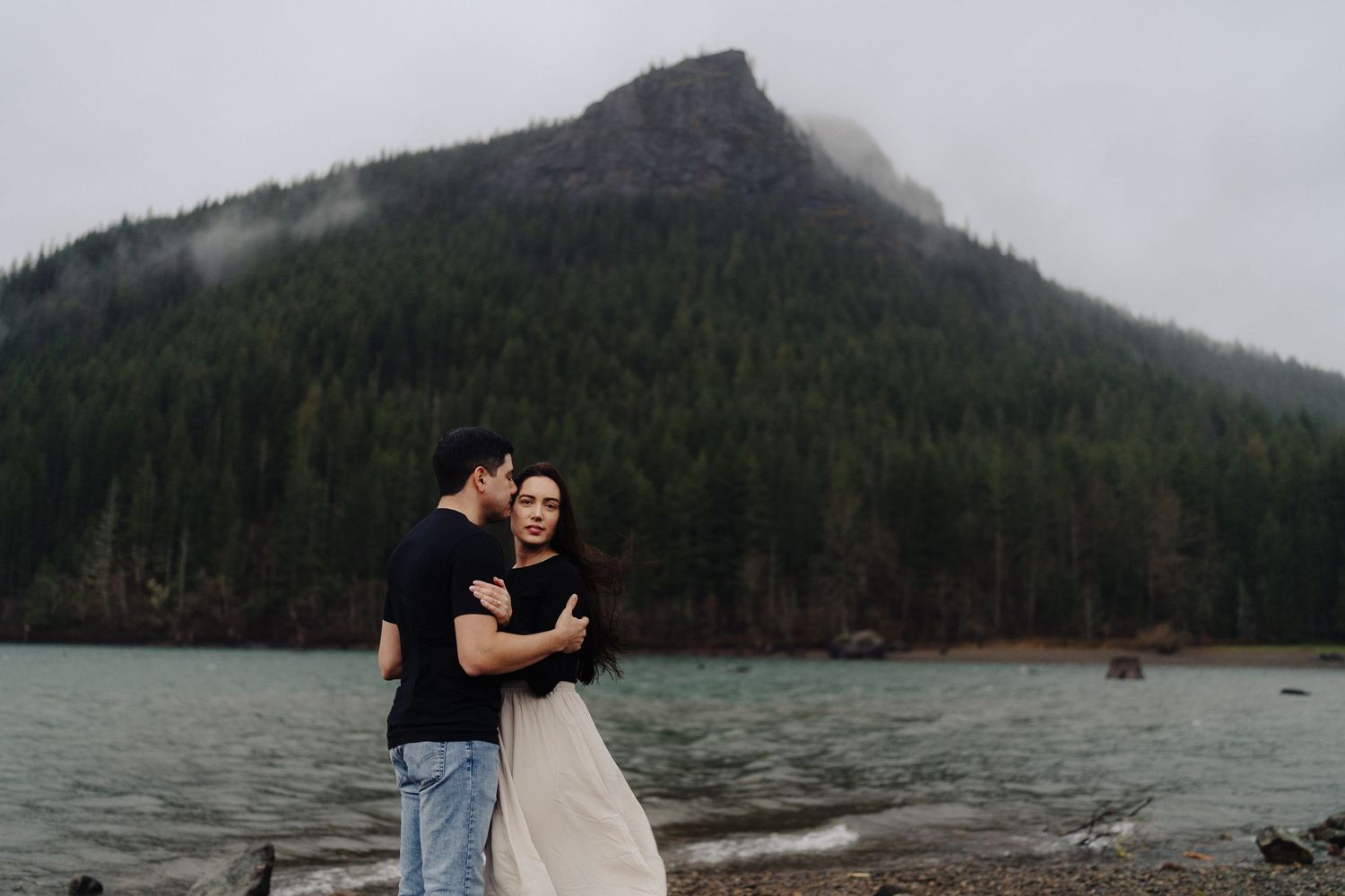 Two people embrace by a misty mountain lake surrounded by evergreen forest on an overcast day.