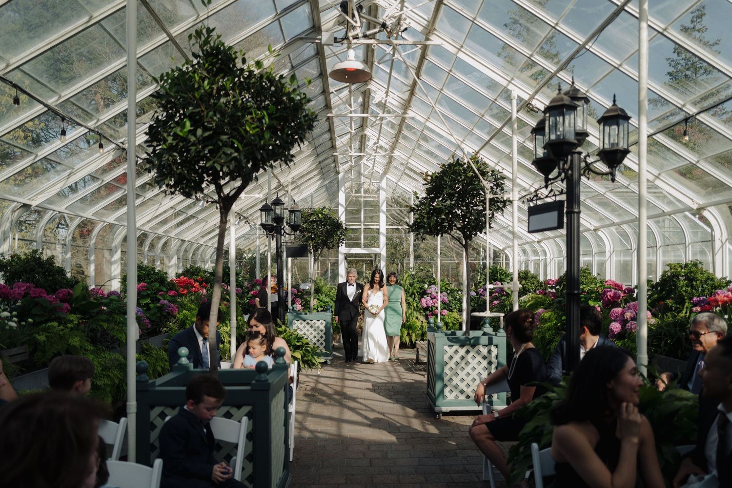 A beautiful greenhouse wedding ceremony with potted trees and ornate lanterns under a glass ceiling.